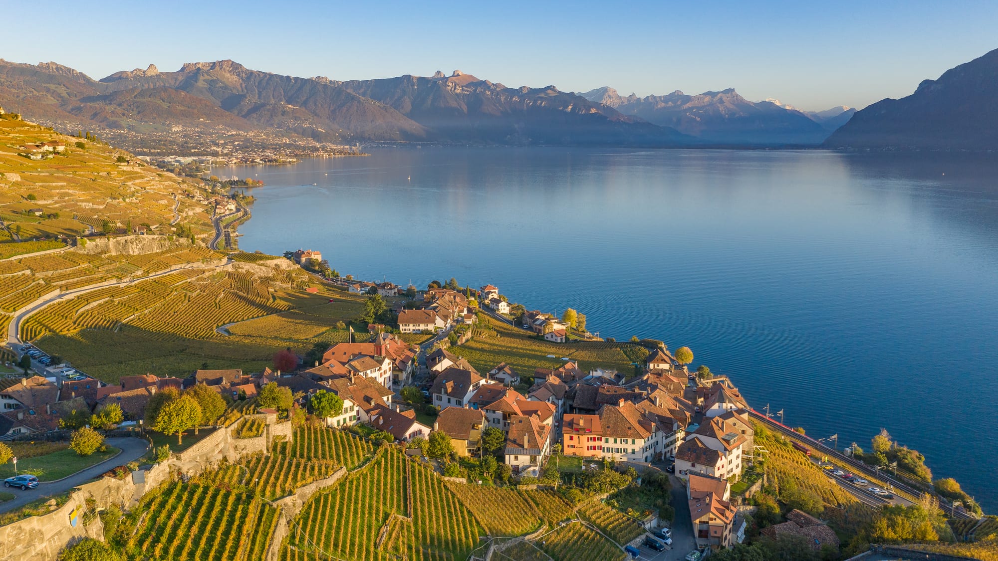 Aerial view of terraced vineyards and a lakeside village of red-roofed houses on Lake Geneva, with mountains beyond