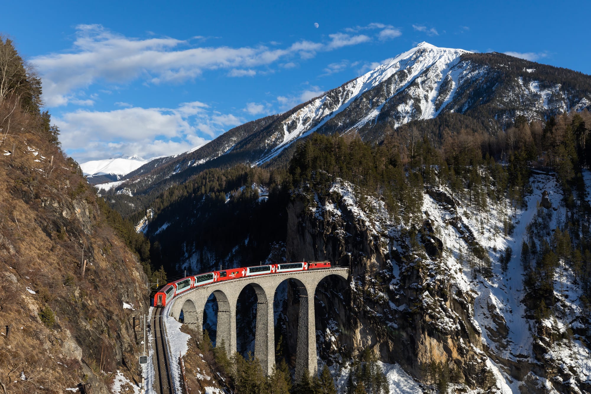 A red train curving across a tall stone arched viaduct over a rocky gorge, with a snow-streaked mountain behind