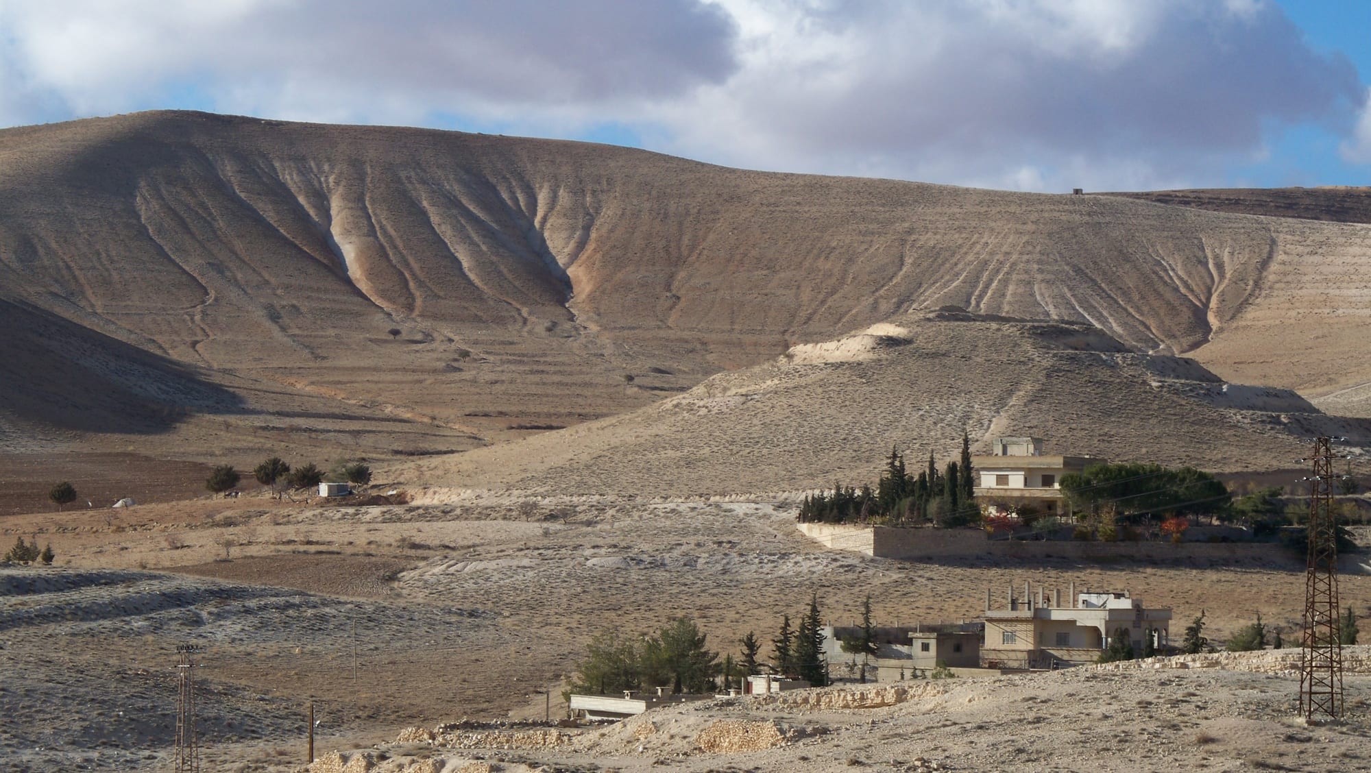 Bare, deeply gullied hills above a few houses and dark cypress trees near Maaloula under a cloudy sky