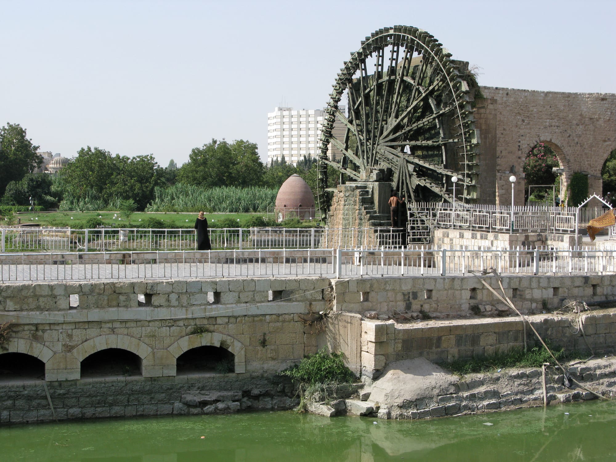 A giant wooden water wheel beside an arched stone aqueduct on the bank of a green river, with gardens and a white tower block behind