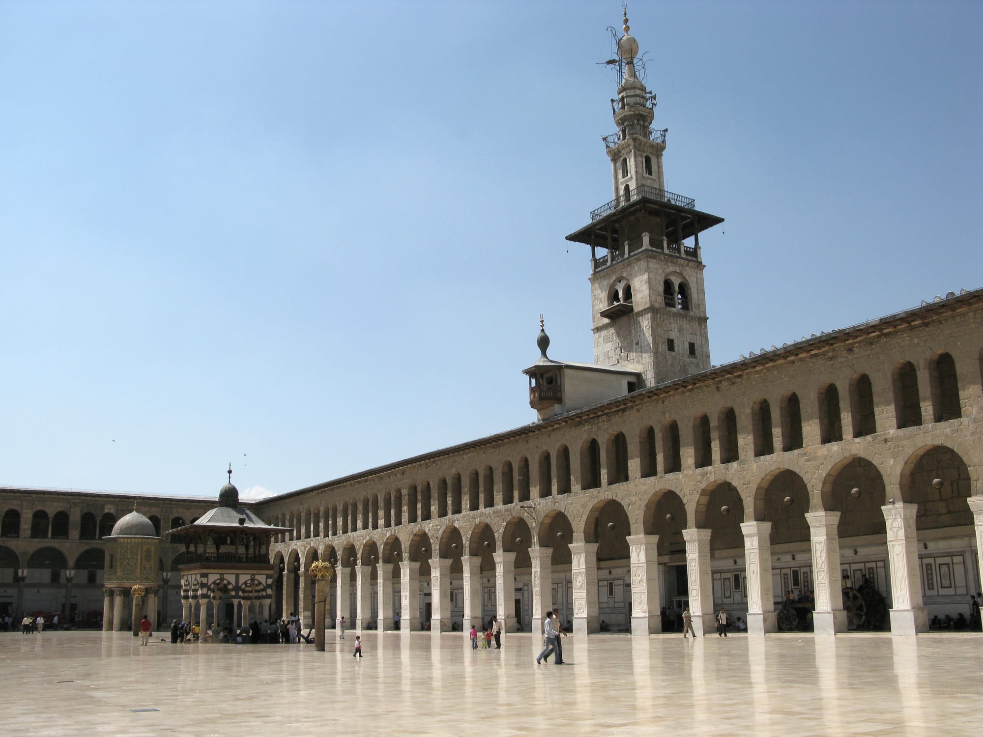 A vast paved courtyard flanked by an arcaded prayer hall with gold mosaic panels, a domed treasury on columns to one side