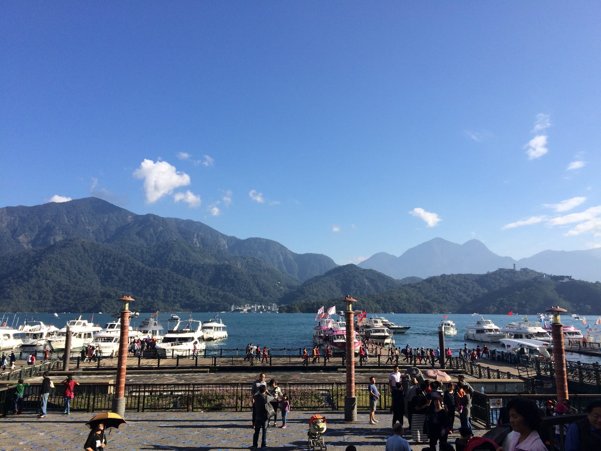Tour boats moored at a lakeside pier, with blue water and forested mountains under a clear sky