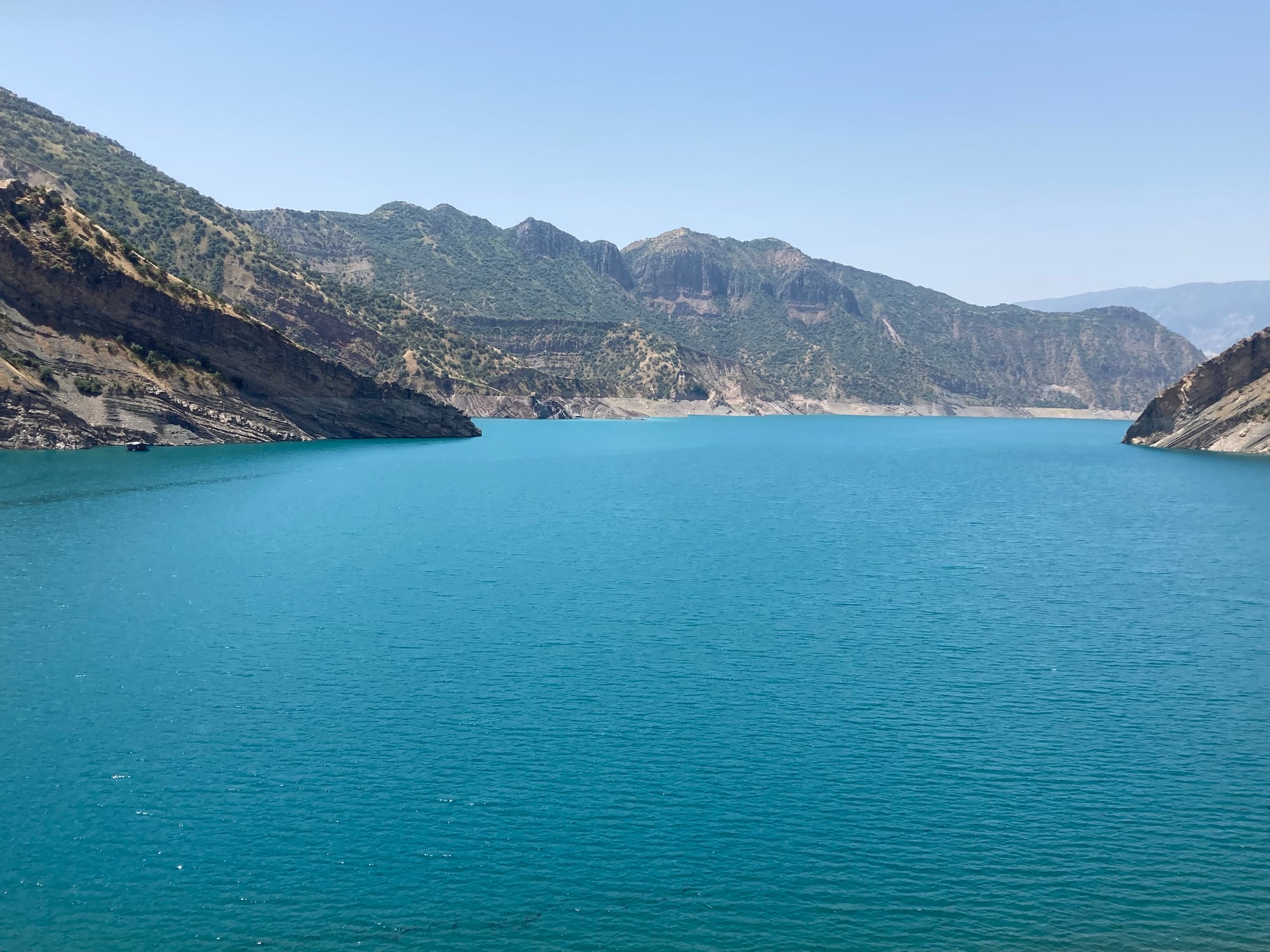 A wide turquoise reservoir filling a valley between dry, scrub-covered mountain ridges under a clear sky