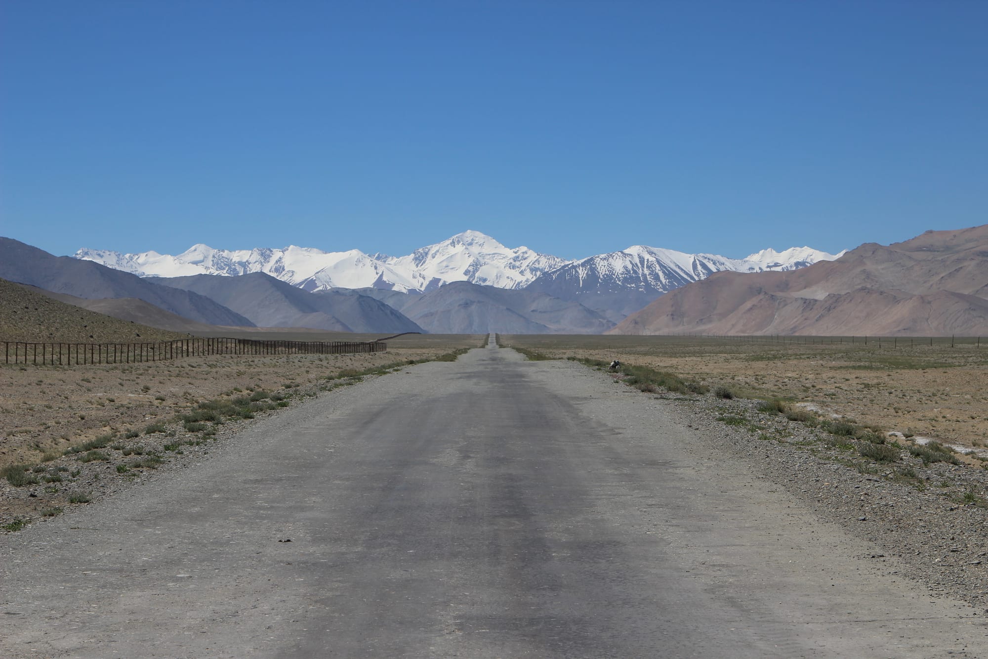 A straight grey road crossing a dry high plain towards snow-covered peaks, with a long fence on the left