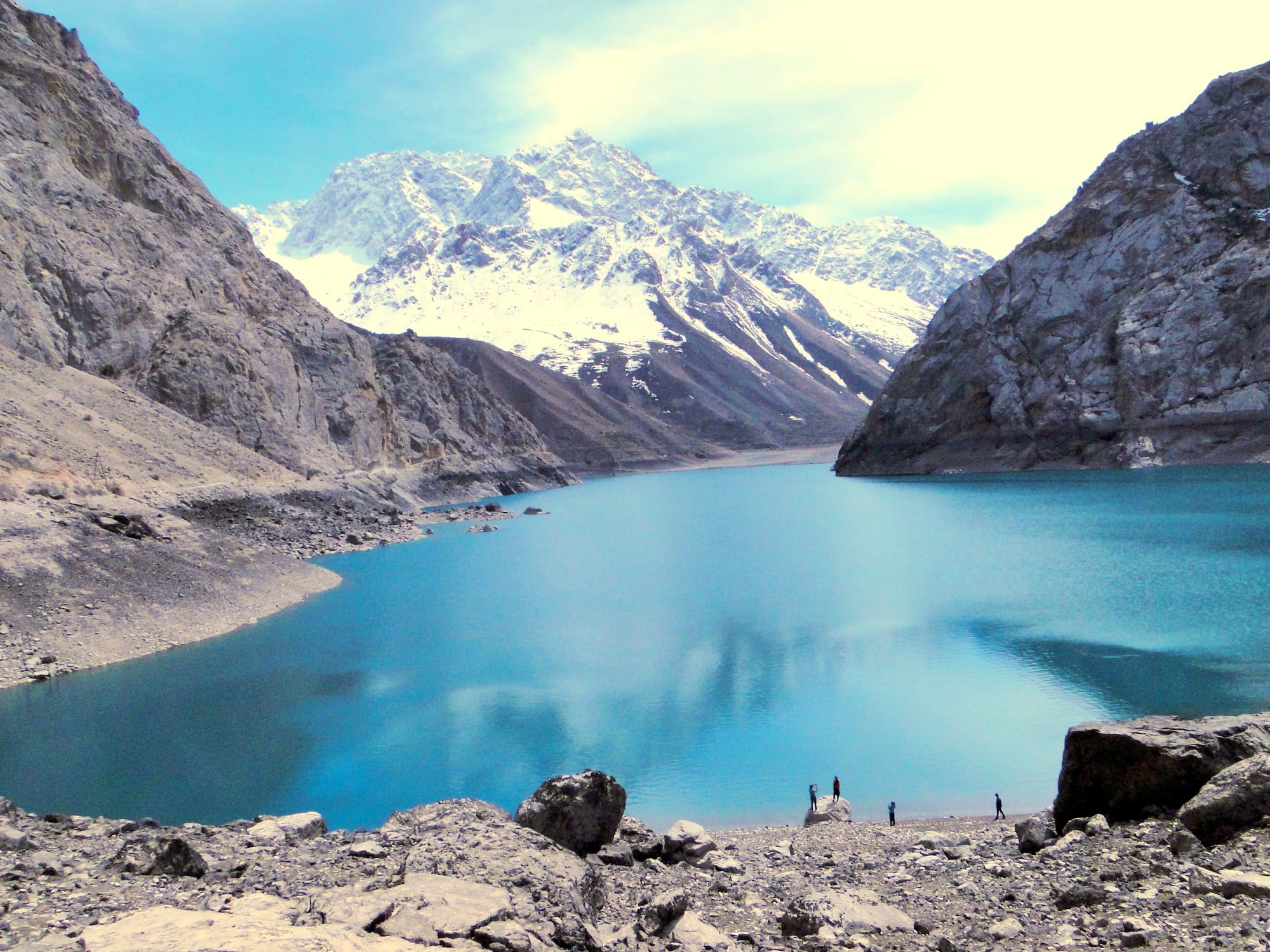 A turquoise lake between grey rocky slopes with a snow-covered mountain behind and a few small figures on the shore