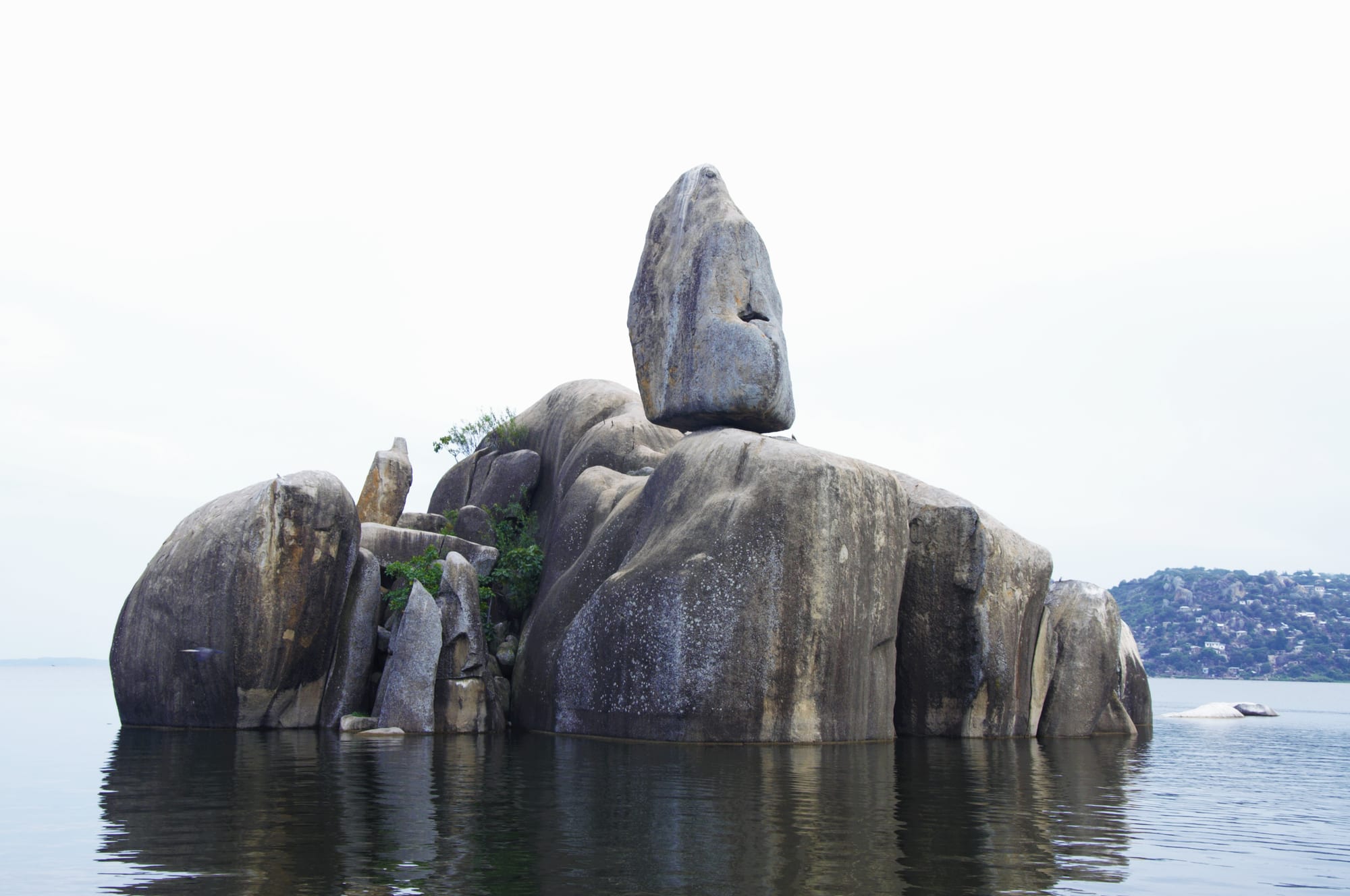 A pile of huge rounded granite boulders with a tall balanced rock on top, rising from calm lake water under a pale sky