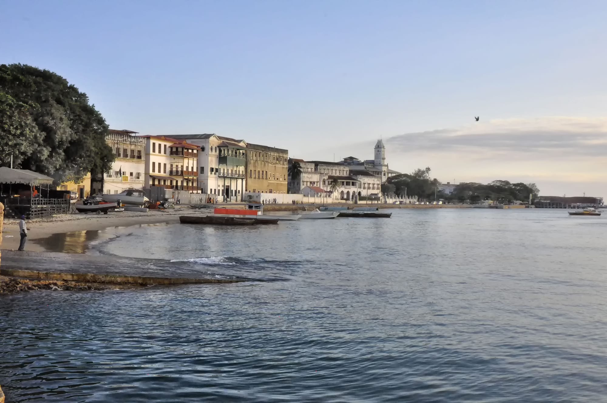 Calm blue water and a sandy shore with moored boats, in front of a row of old multi-storey buildings and a white tower