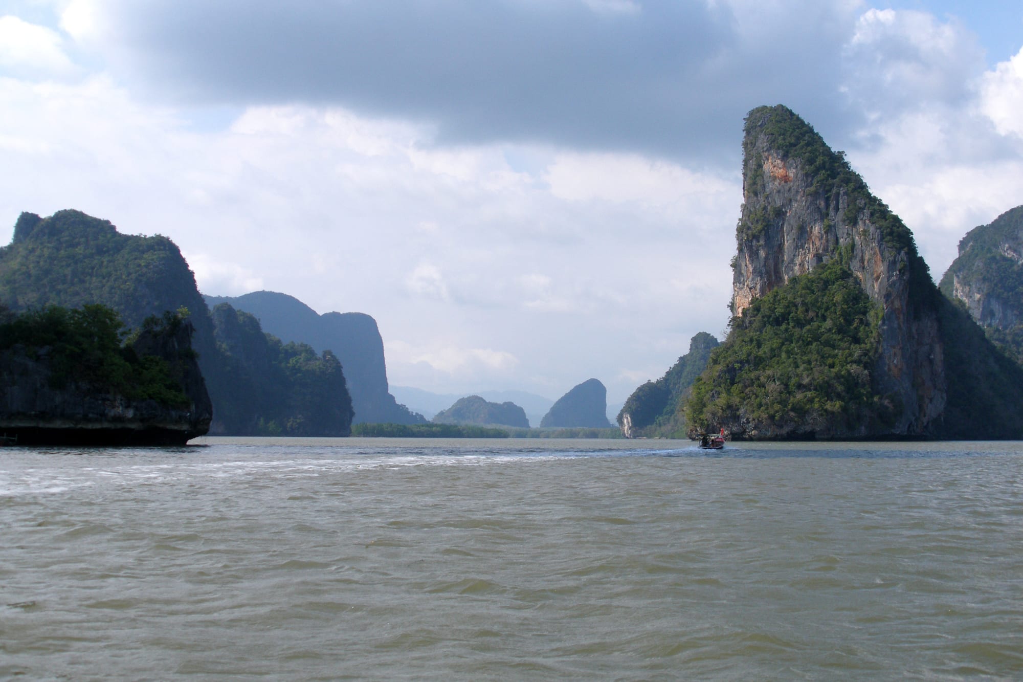 Sheer, forest-topped limestone towers rising from grey-green water, with a small longtail boat passing beneath them