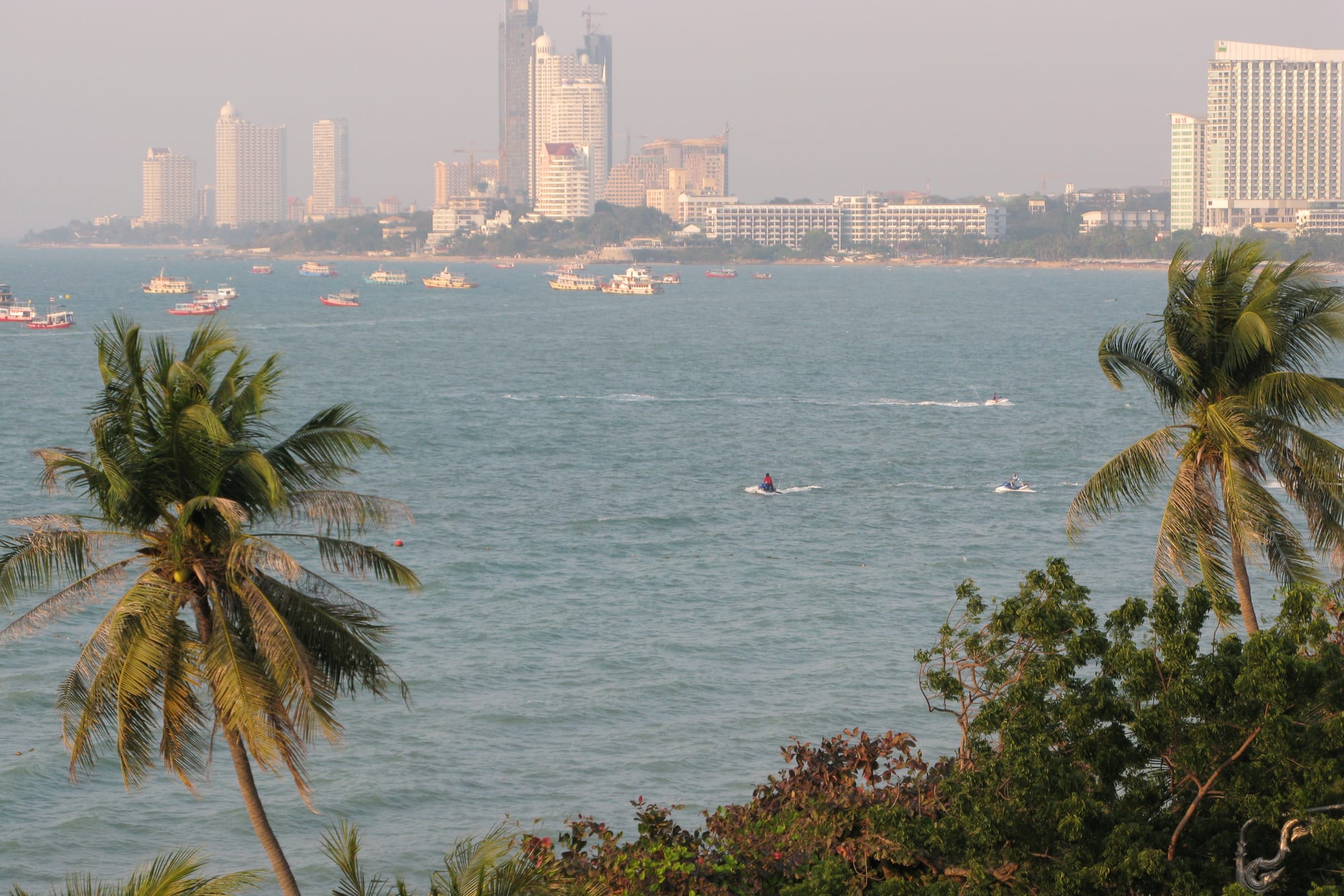 Palm trees framing a wide blue bay dotted with boats and jet skis, with high-rise hotels along the far shore