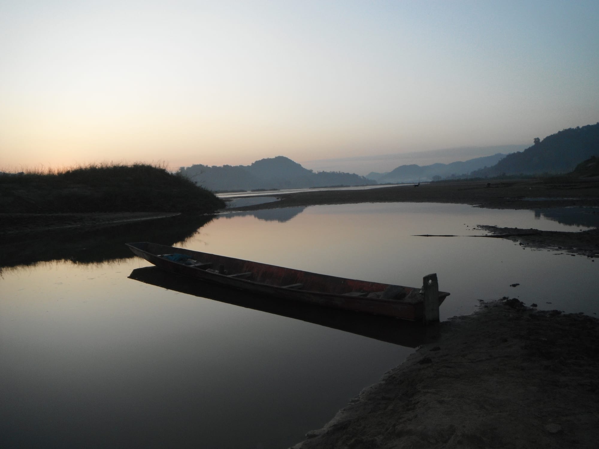 A wooden longboat moored in still water on the Mekong at dusk, with hazy forested hills across the river