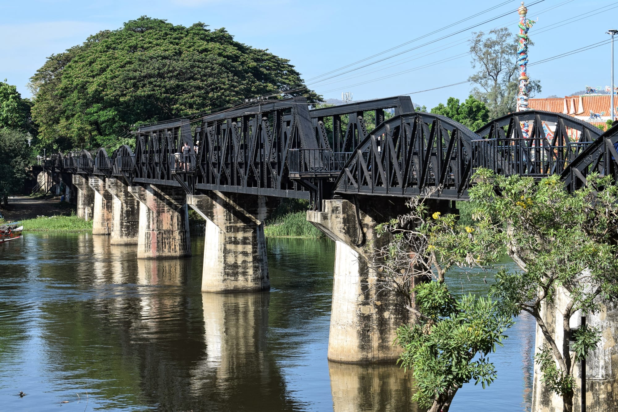 A black steel railway bridge with curved trusses on concrete piers, crossing a calm brown river lined with trees