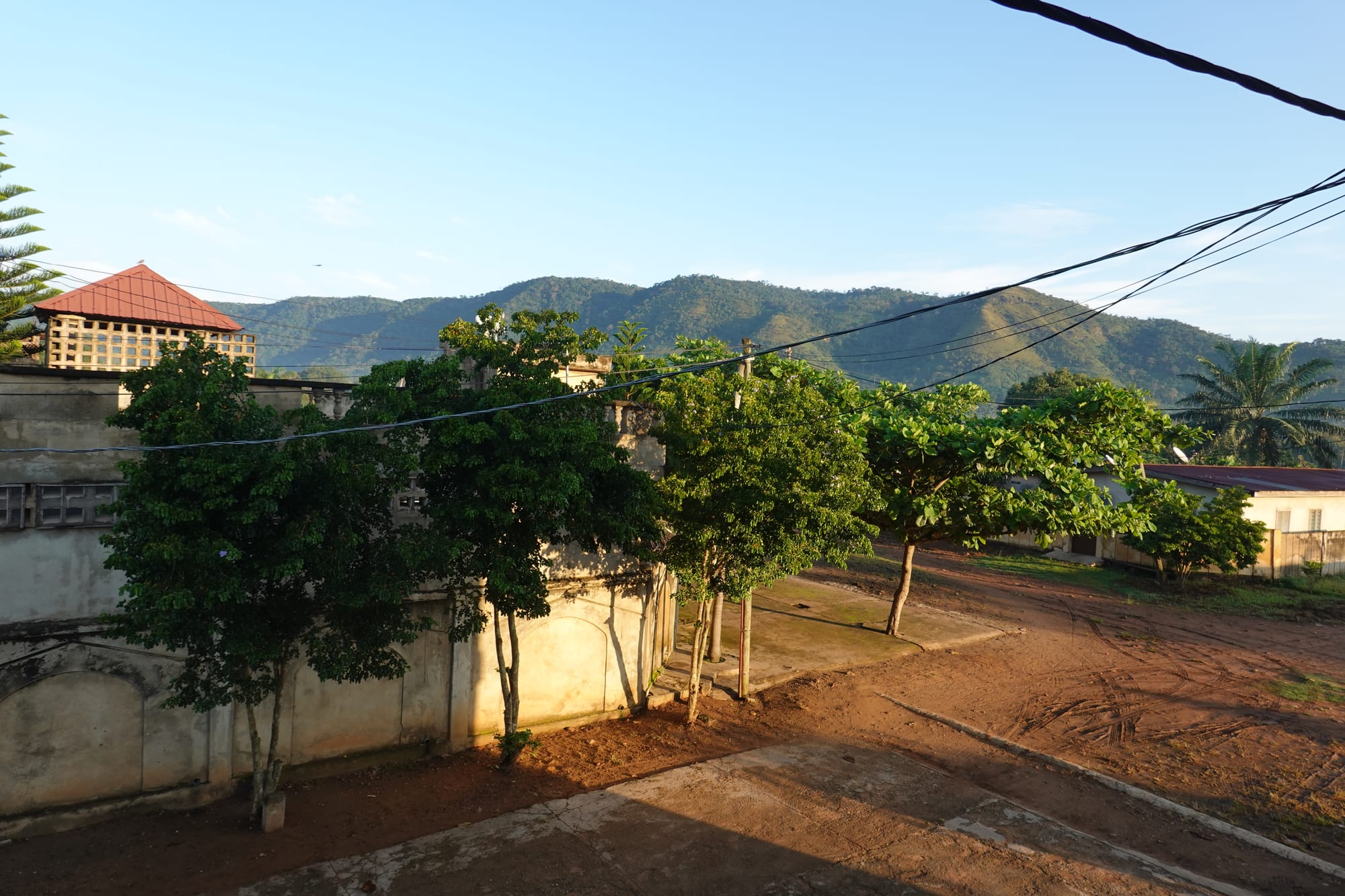Green trees and houses along a red-earth street, with a range of wooded mountains behind under a blue sky