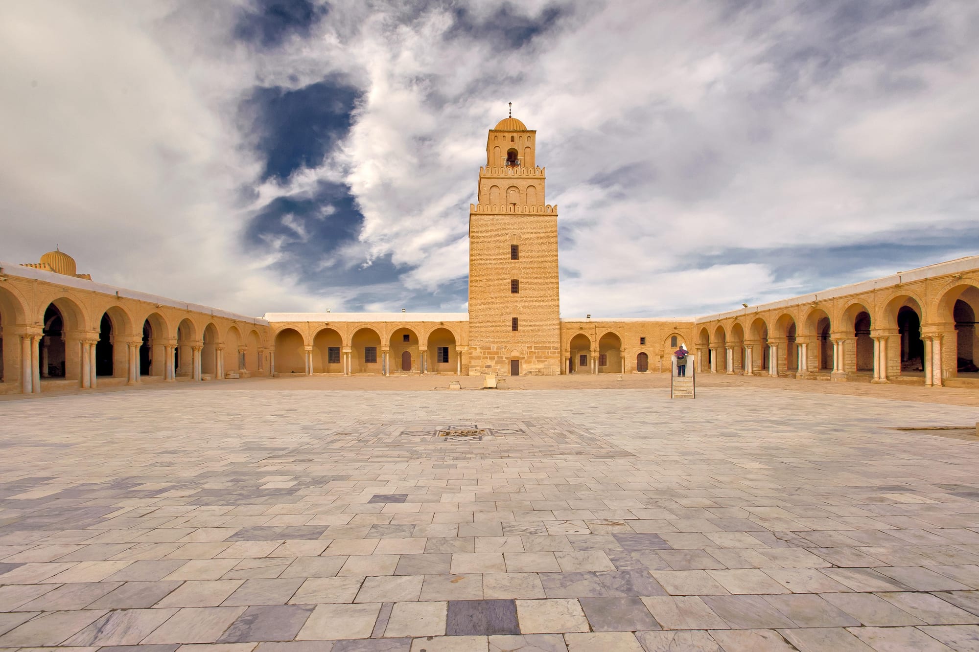 A wide marble-paved courtyard ringed by arcades, with a square stone minaret under a cloudy sky