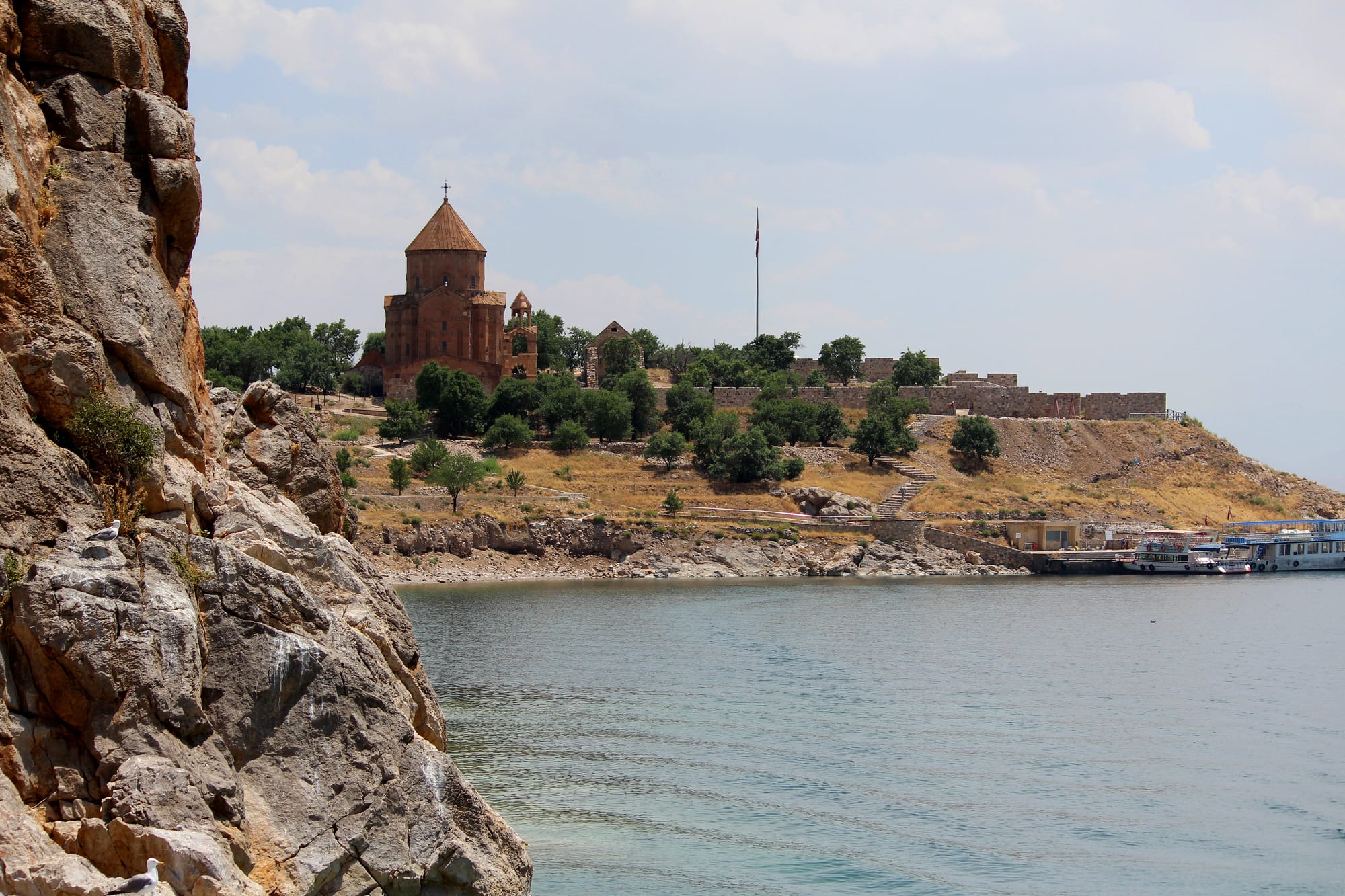 A domed red-stone church on a dry island hillside above calm blue lake water, seen past a rocky cliff
