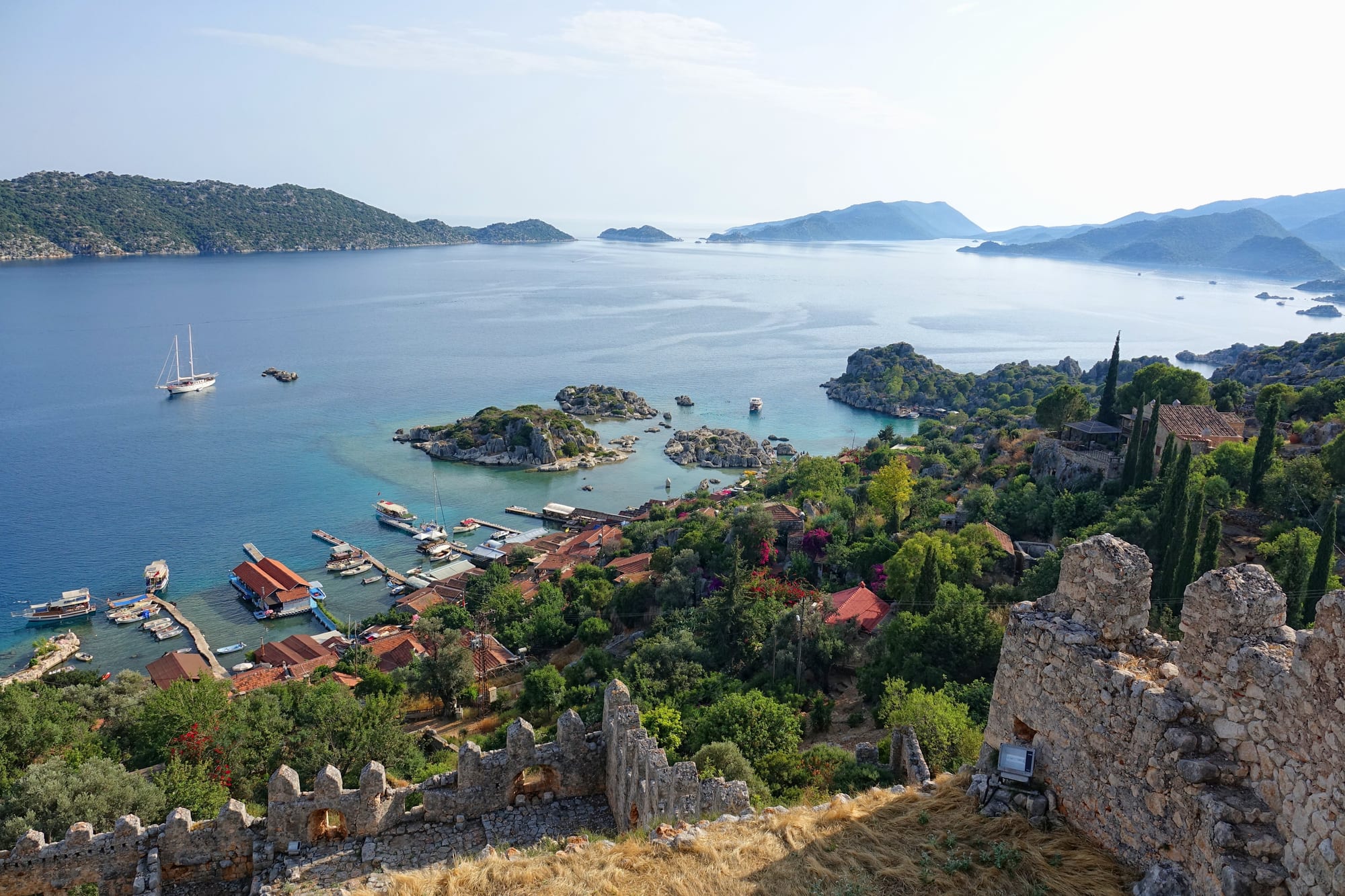 Medieval stone battlements above a village of red roofs, with boats moored in a calm blue bay among green islands