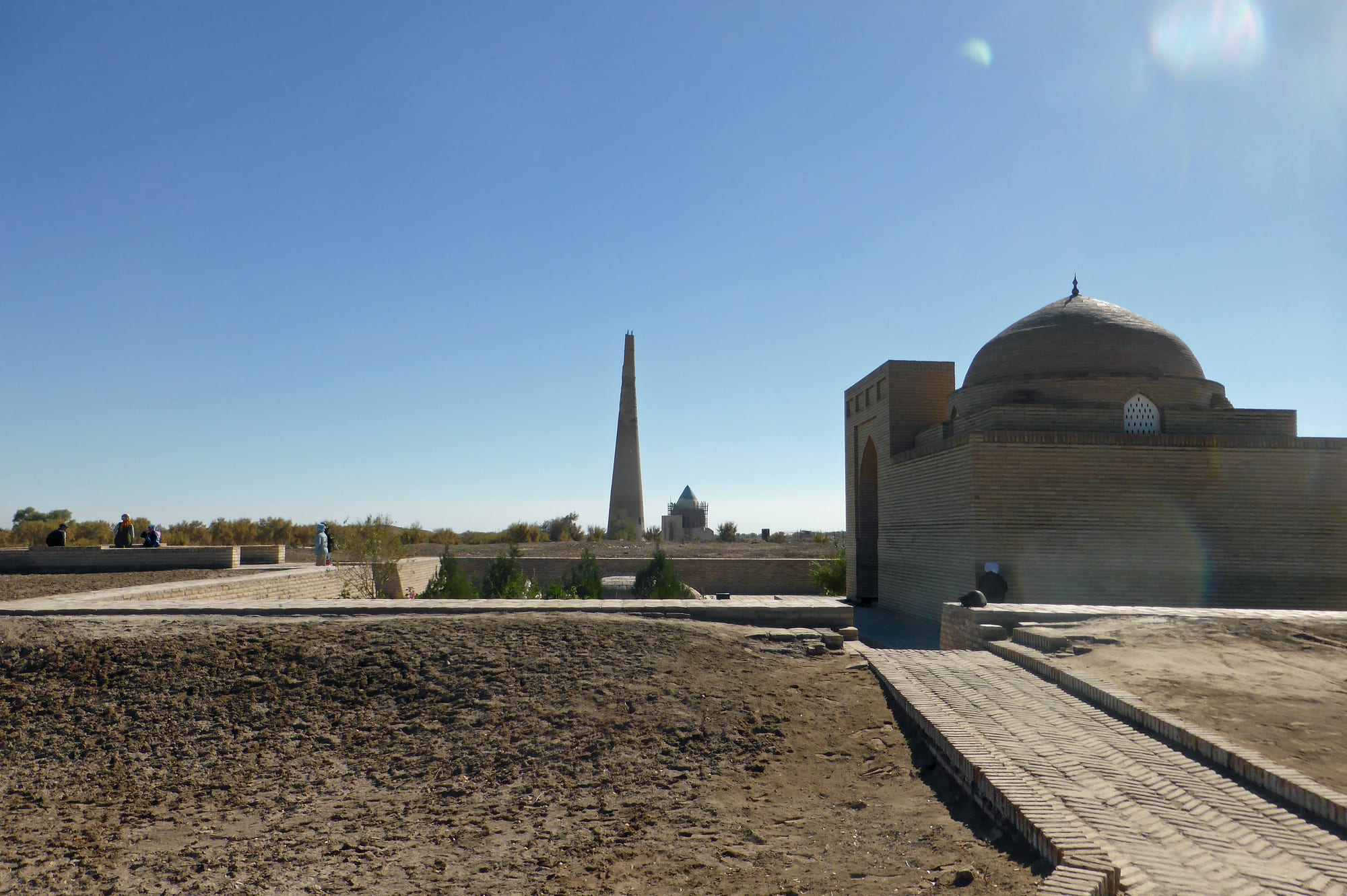 A domed brick building and paved paths on dry ground, with a tall slender minaret and a small conical-domed tomb in the distance under a clear sky