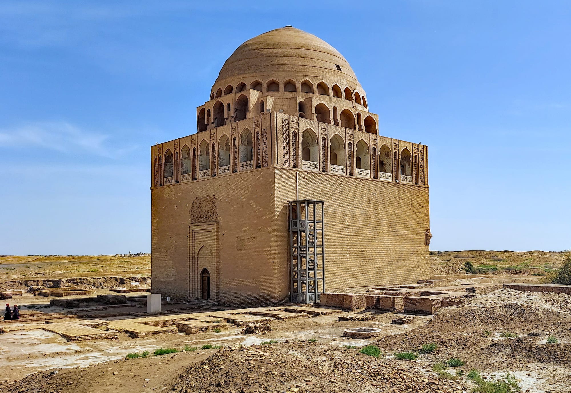 A square brick mausoleum with an arcaded gallery and a large ribbed dome, standing among low excavated ruins