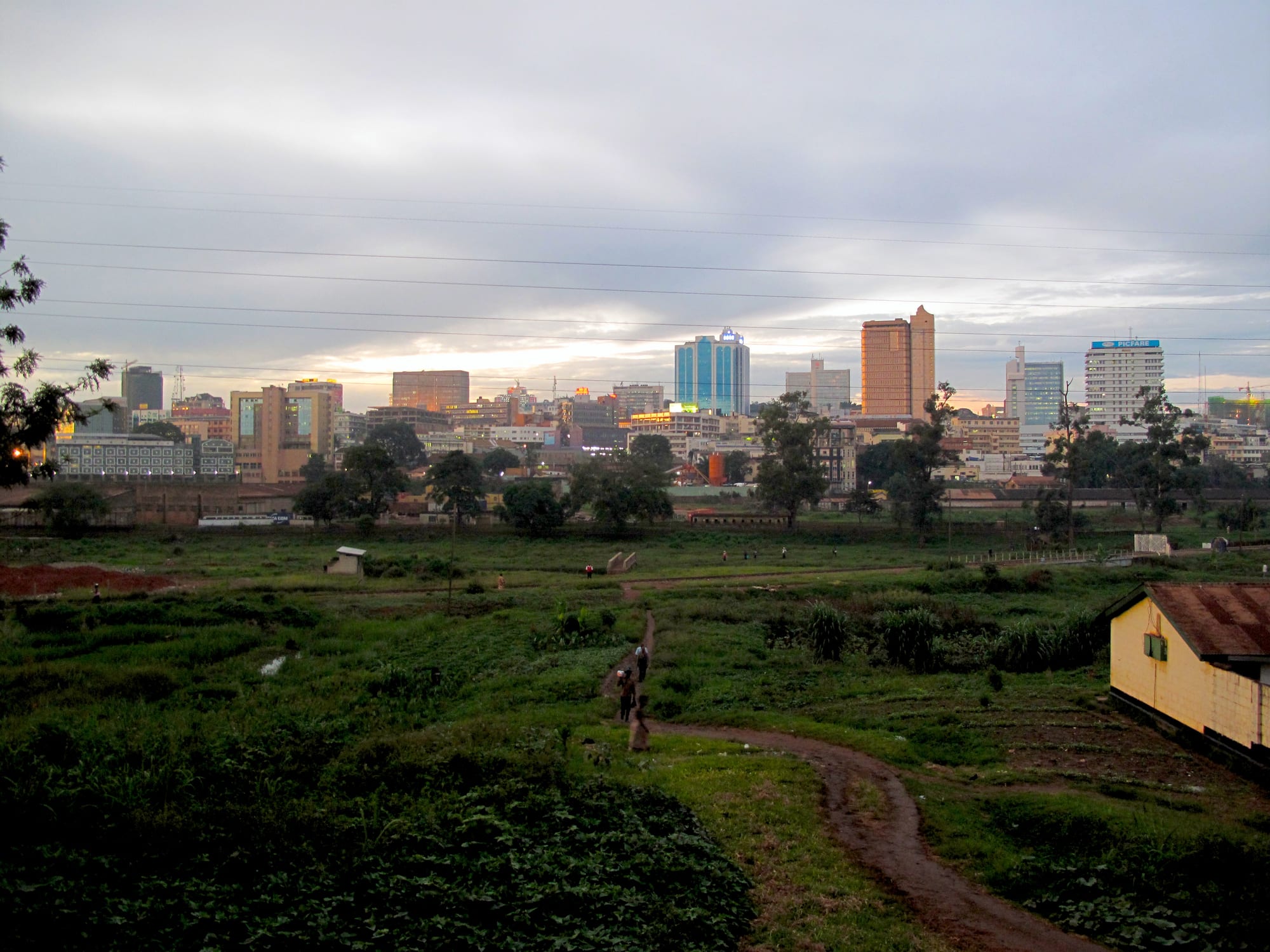 Kampala's high-rise centre at dusk seen across green open ground, with a dirt path and figures walking in the foreground
