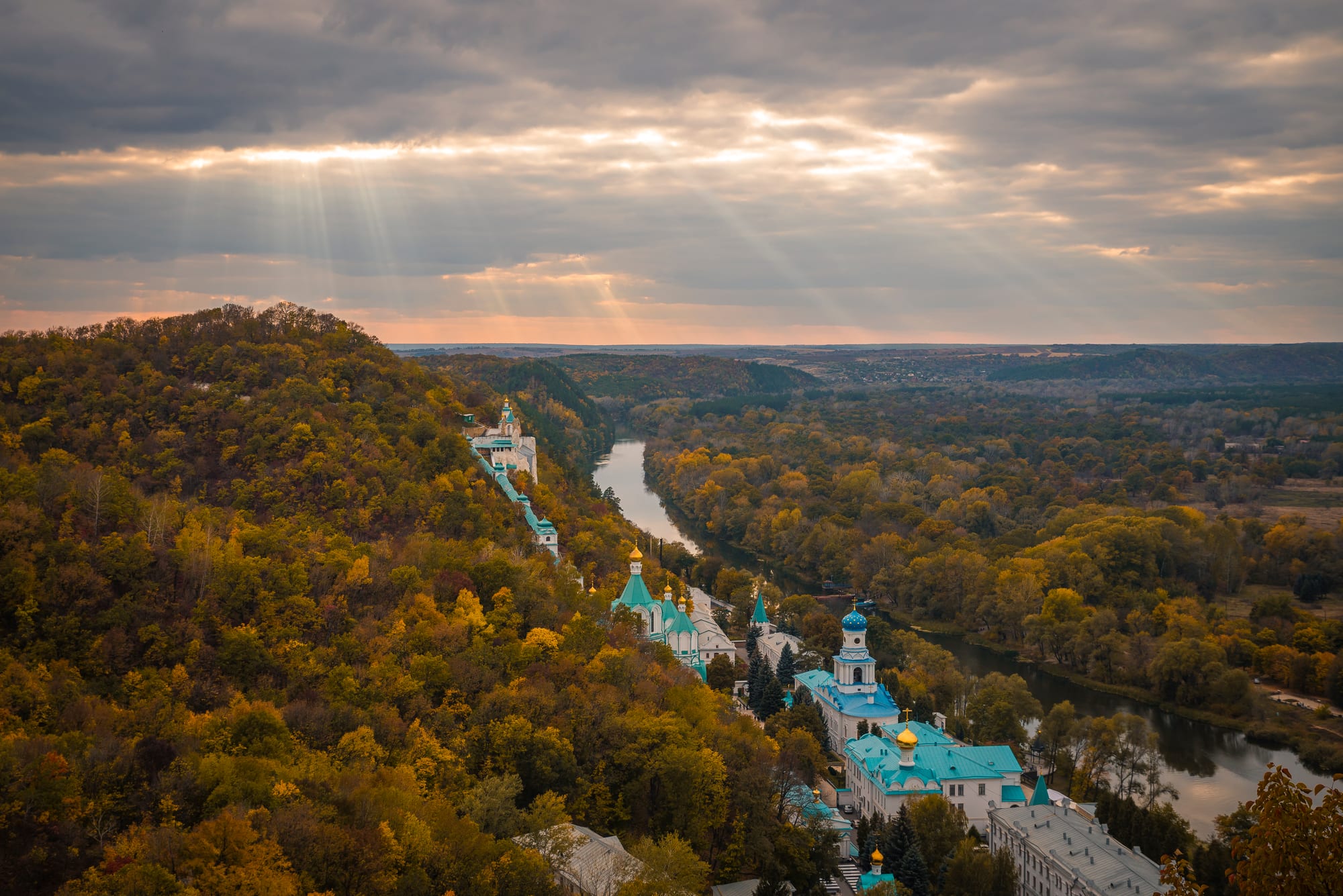 Monastery churches with blue and gold domes on a wooded hillside in autumn colours above a winding river, under sunbeams
