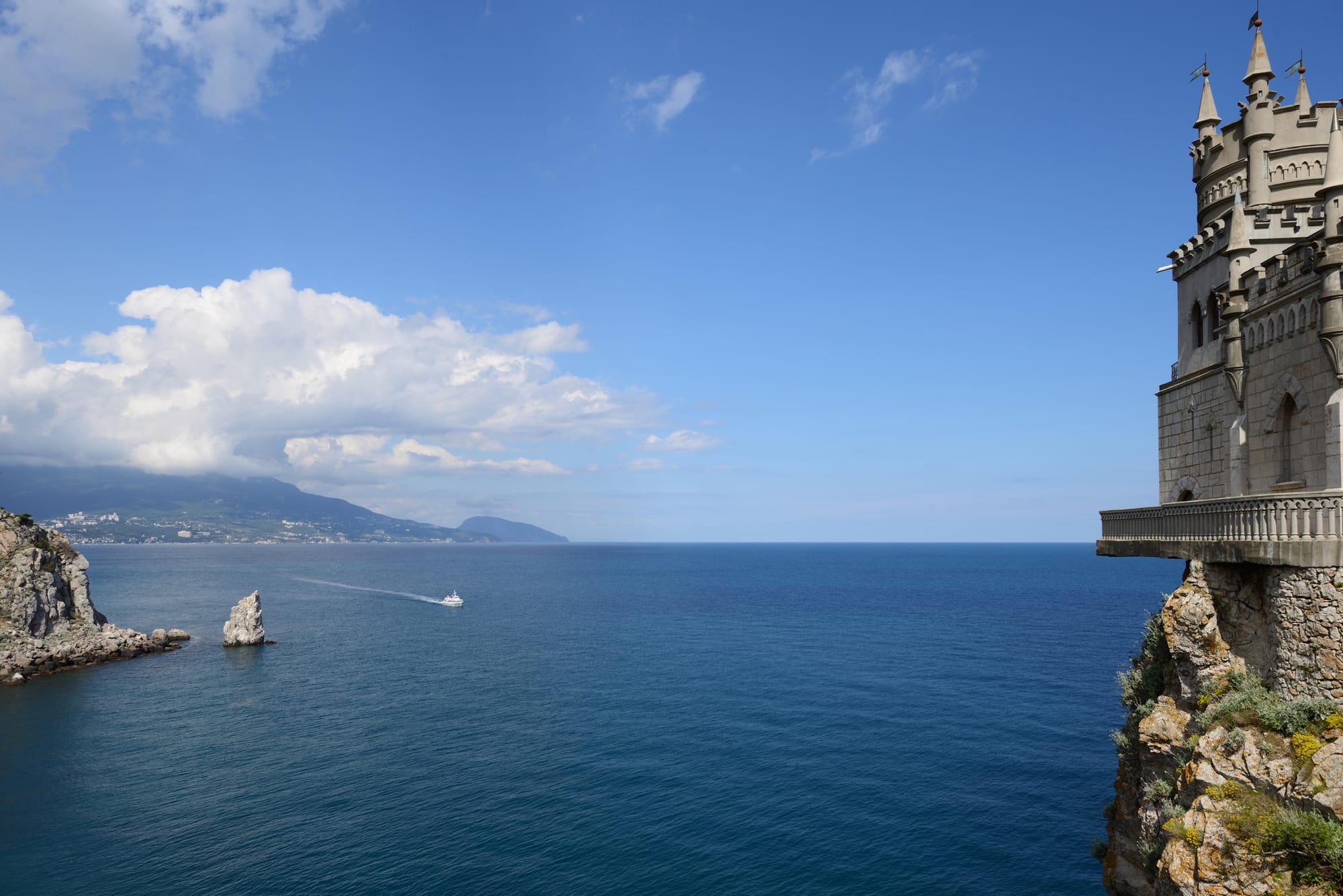 A small turreted castle on a cliff edge above a deep blue sea, with a boat, a rock stack and wooded mountains across the bay