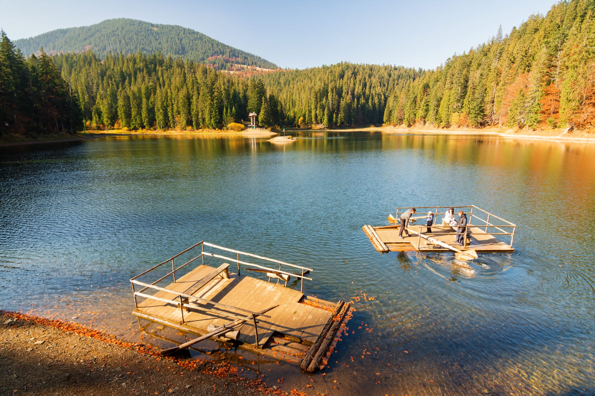 A clear mountain lake ringed by dense fir forest in autumn, with two wooden rafts on the water in the foreground