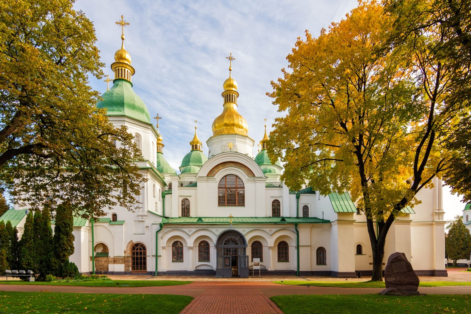 A white cathedral topped with clustered green and gold domes, beside a tall pale bell tower, under a blue sky