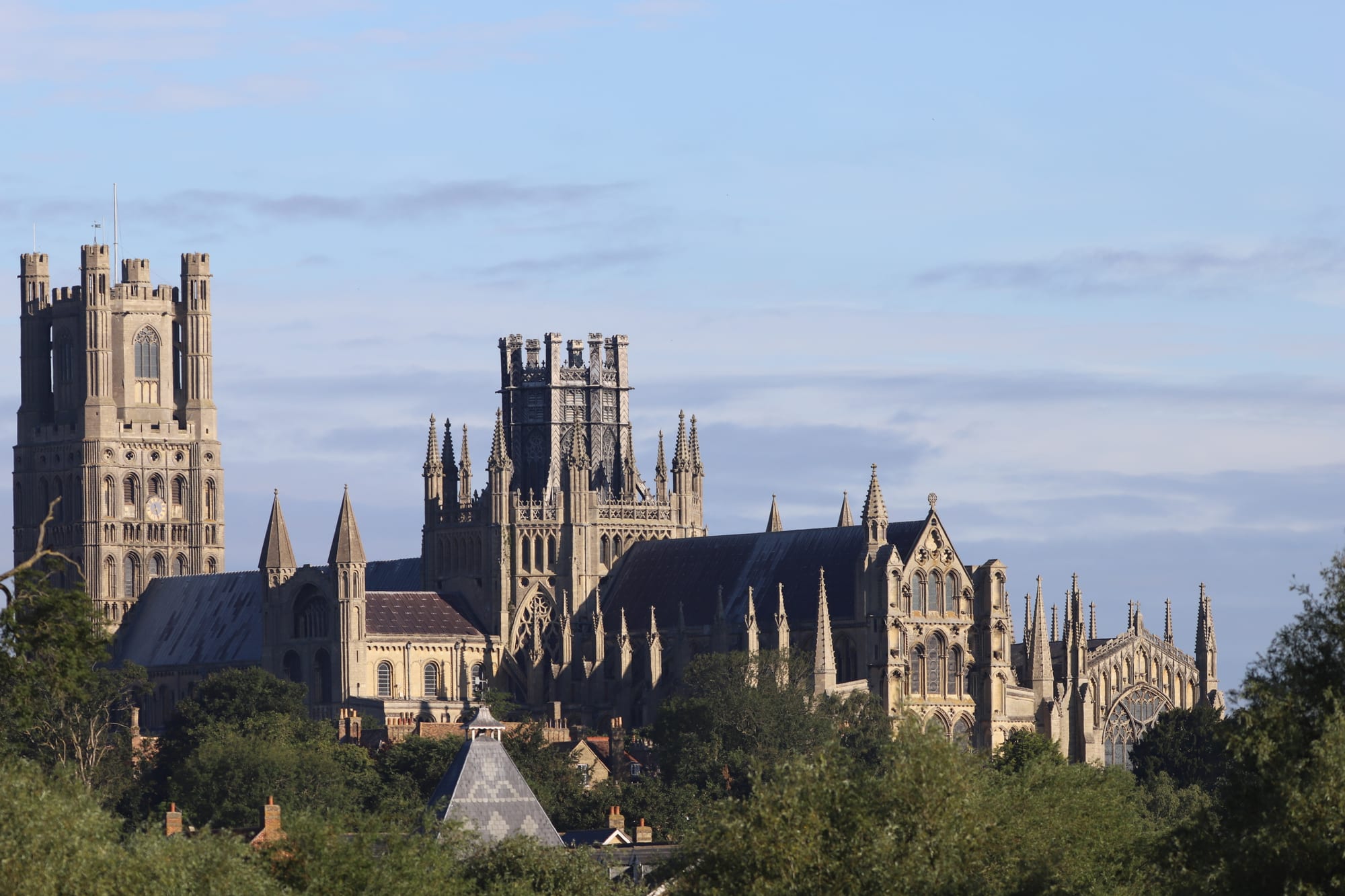 Ely Cathedral's square west tower and lantern-topped octagon rising above trees and rooftops under a pale blue sky