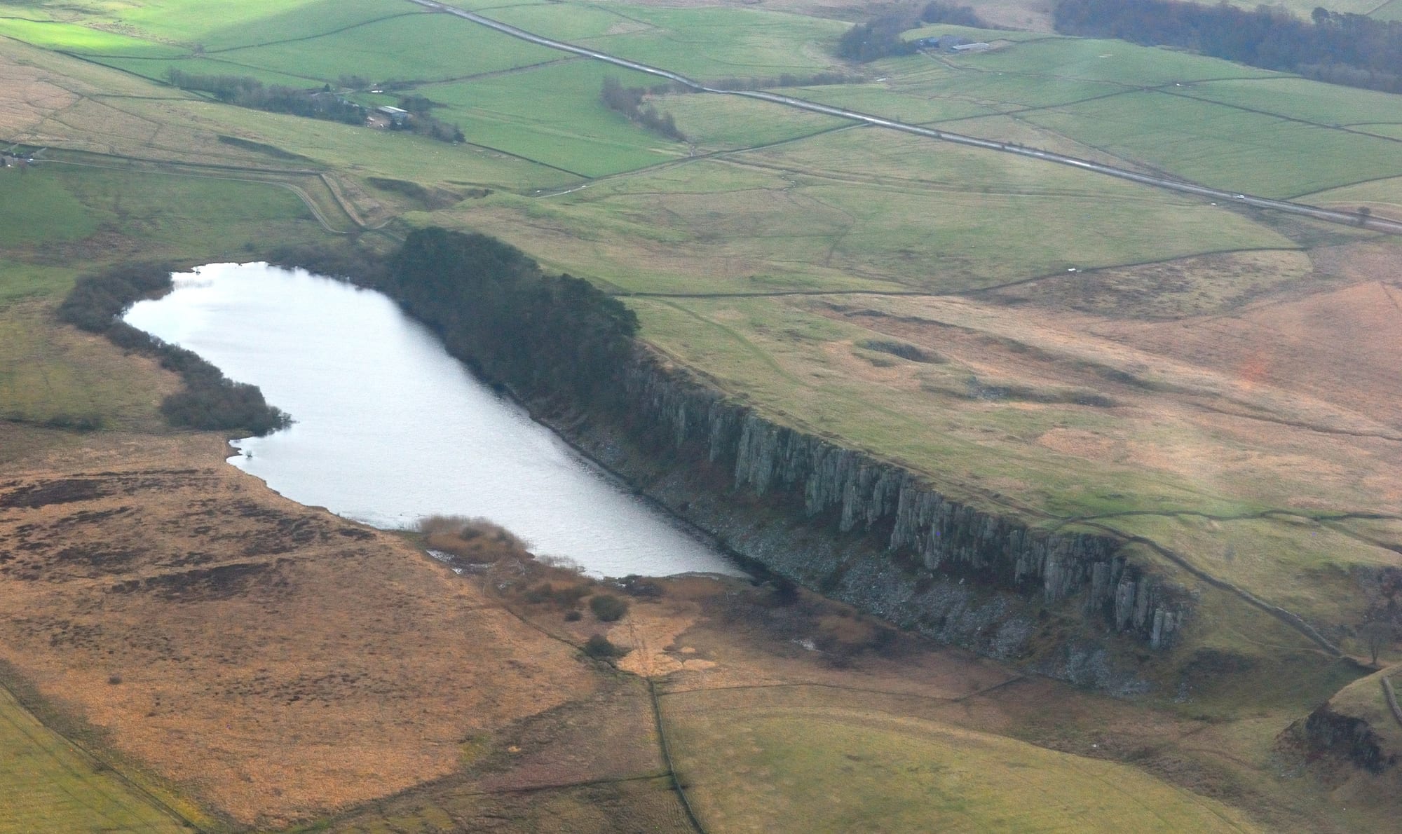 Aerial view of a small grey lake beneath a line of dark cliffs, surrounded by green fields and brown moorland