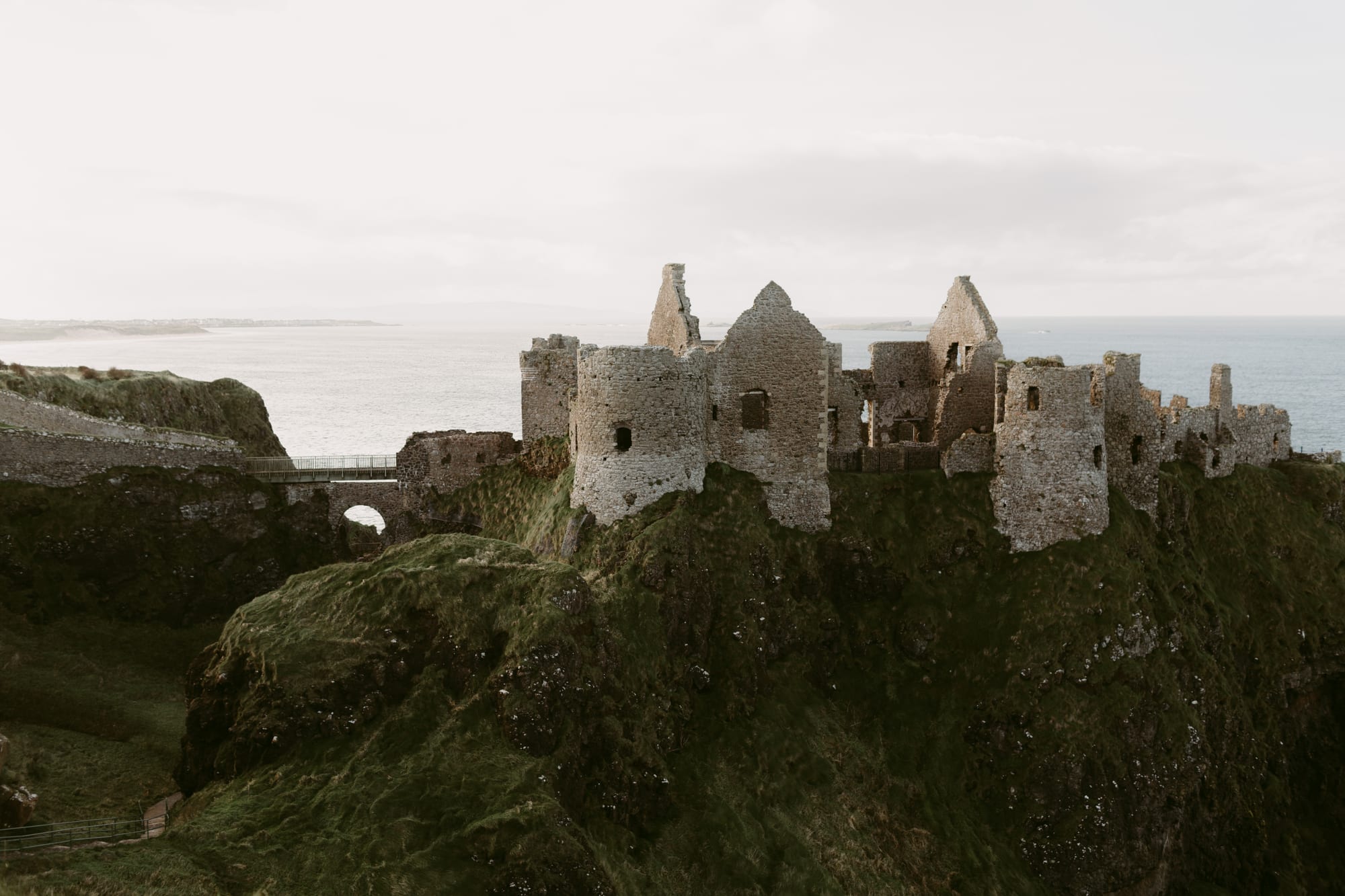 Ruined stone towers and gables of a castle on a grassy clifftop above a pale sea and distant coastline