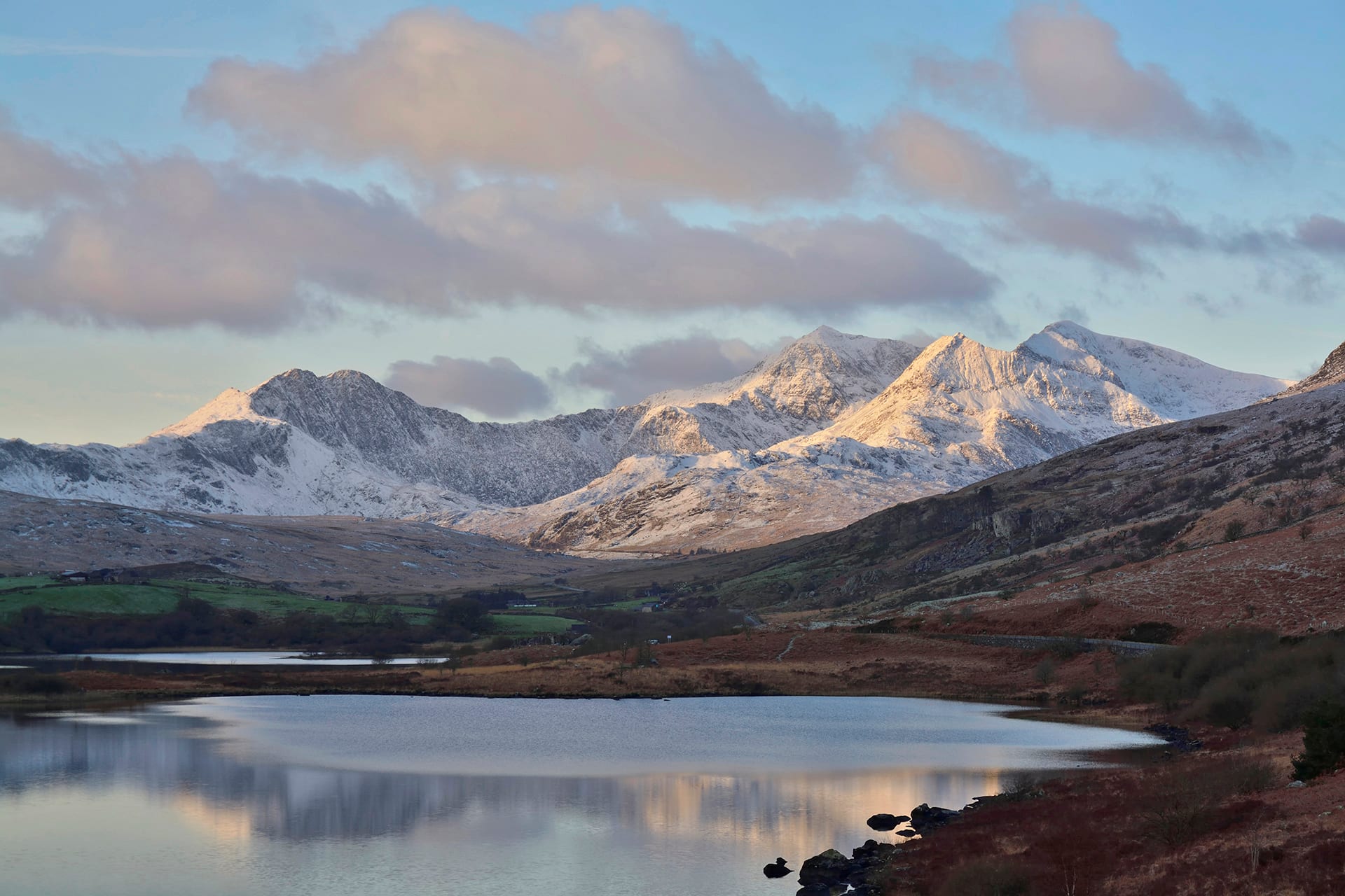 Snow-covered mountain peaks lit by morning sun rising above a calm lake and brown moorland