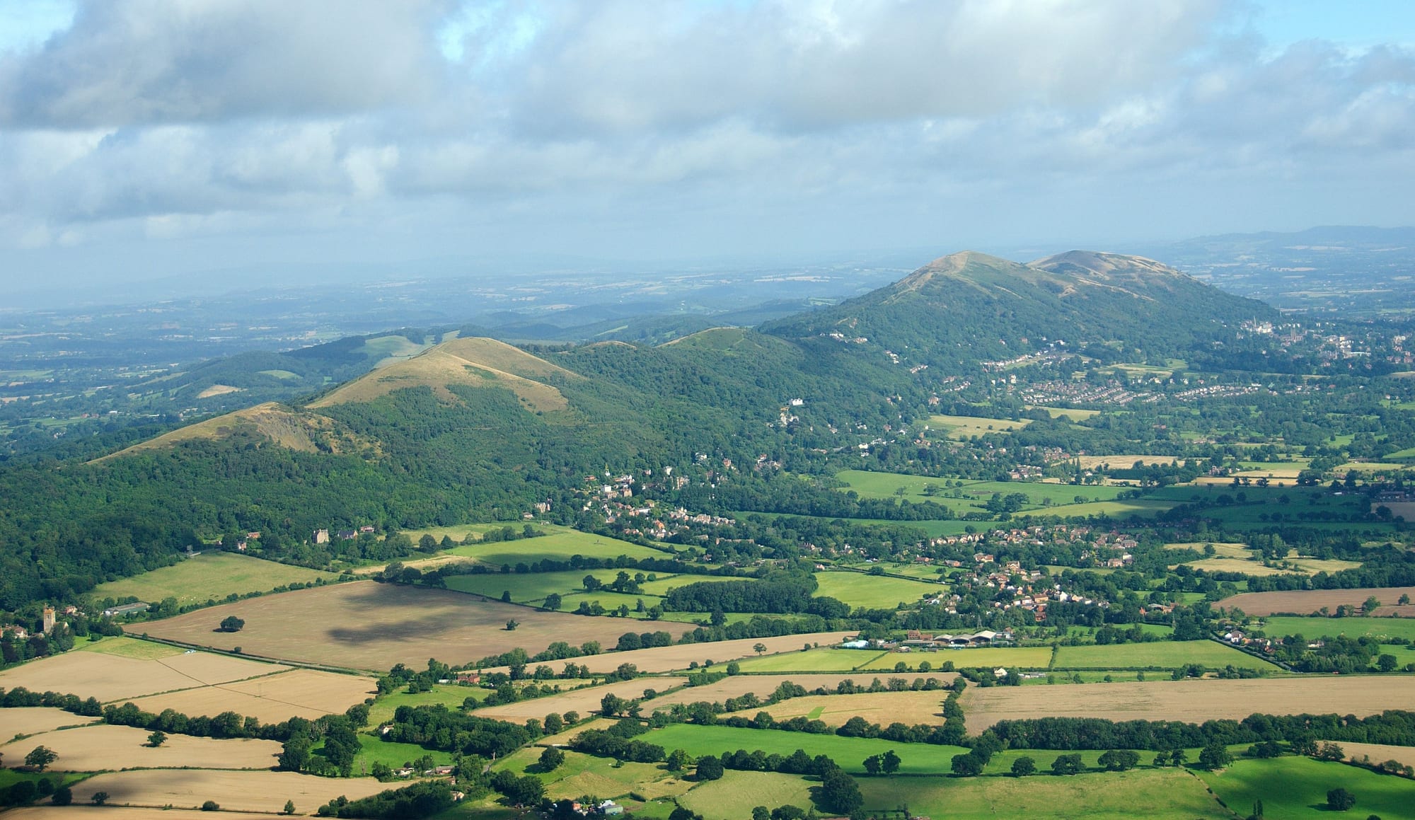 Aerial view of the green, wooded Malvern Hills rising above patchwork fields, villages and the town of Malvern
