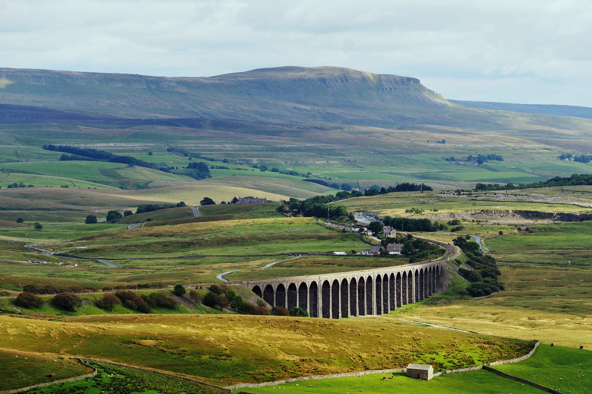 A long stone railway viaduct of many arches crossing green moorland, with the flat-topped hill of Pen-y-ghent behind
