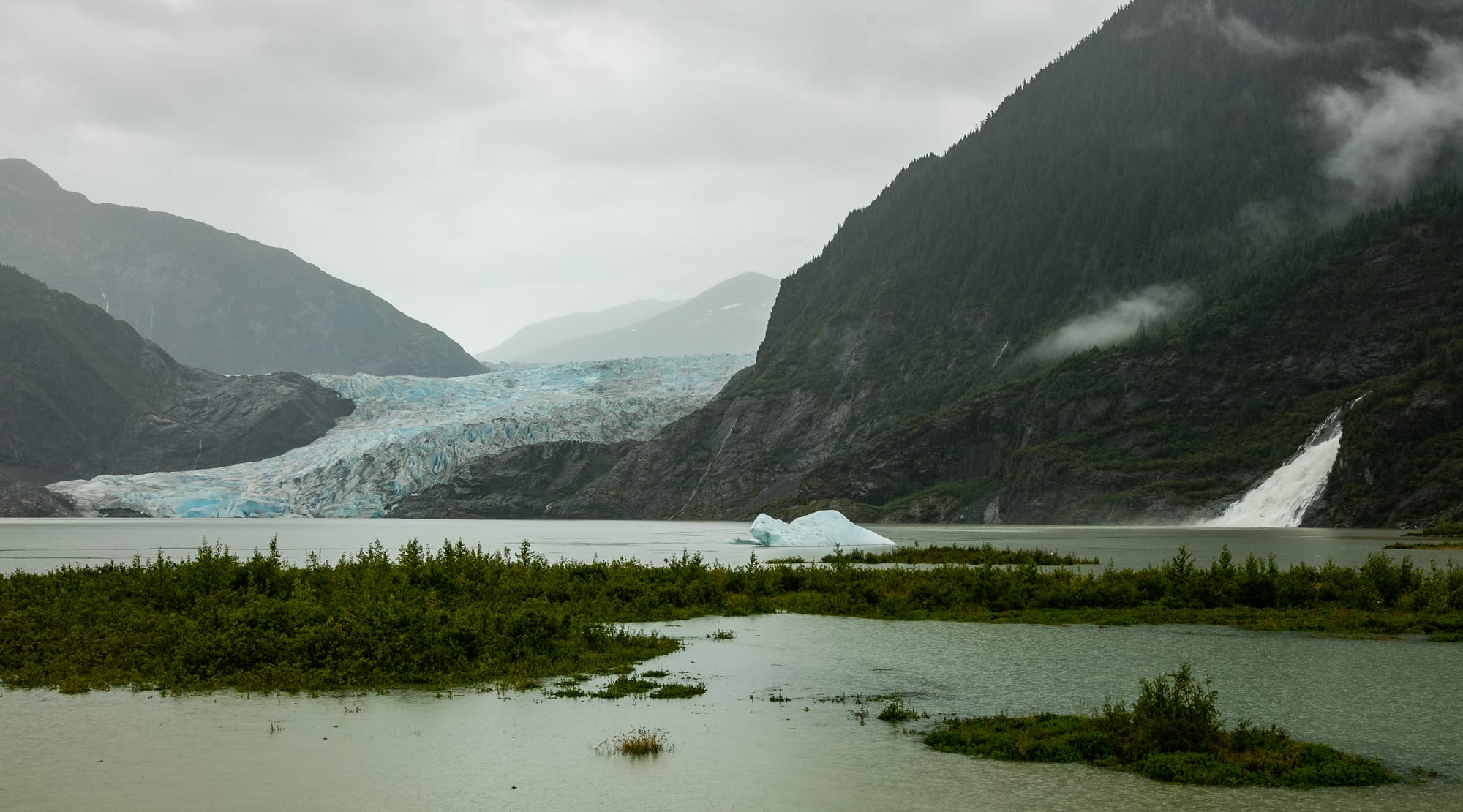 A blue-white glacier flowing into a grey lake between forested mountains, with a waterfall on the right and low cloud