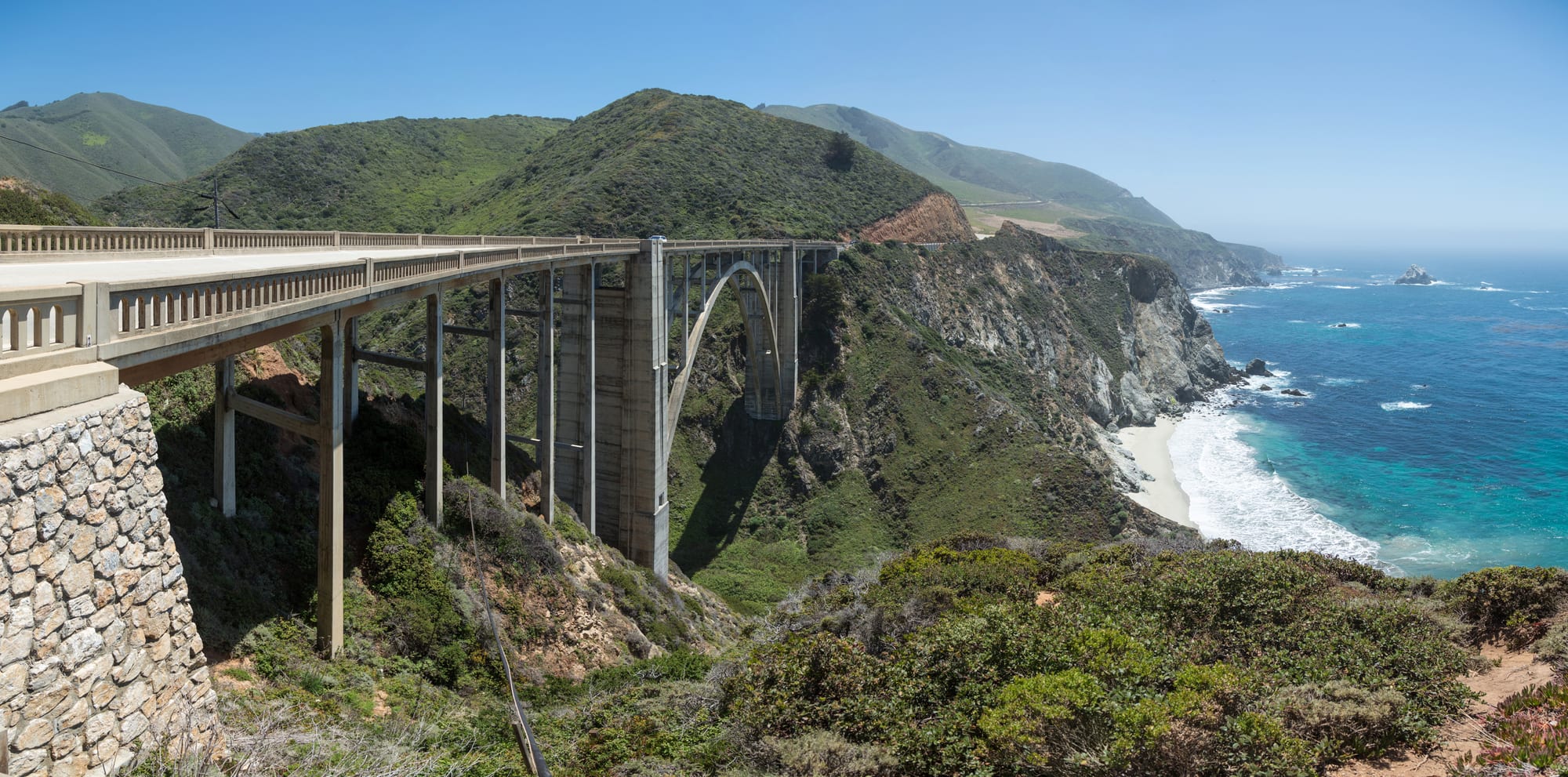 A concrete arch bridge spanning a green canyon above a sandy cove, with steep coastal hills and a turquoise sea behind