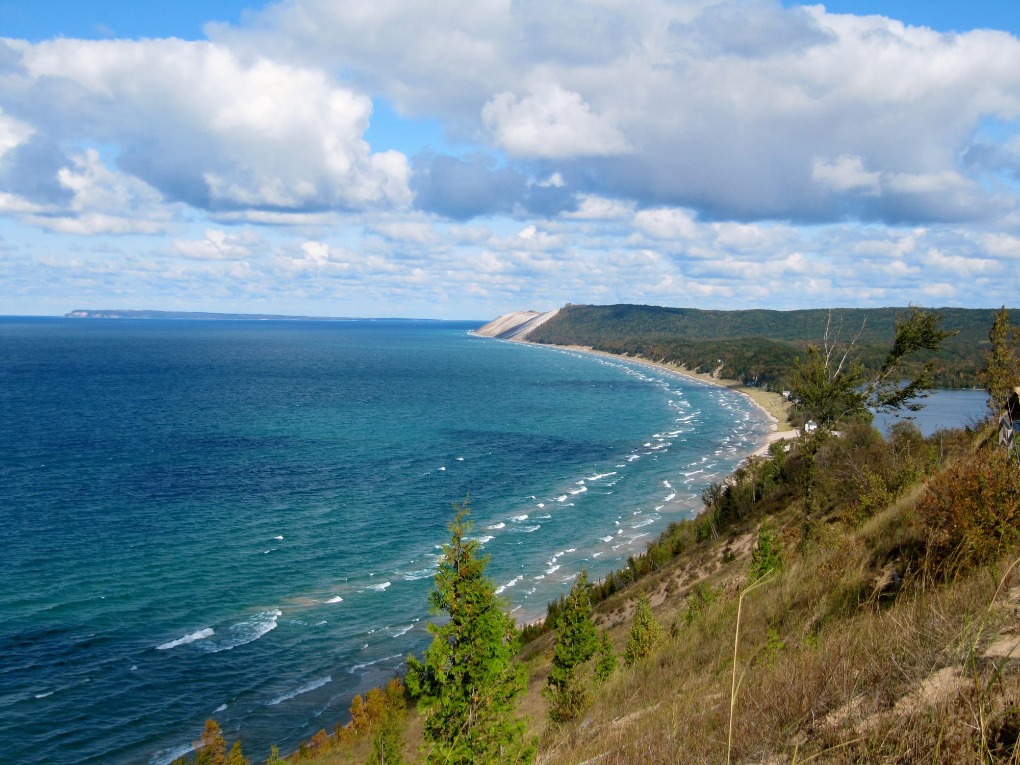 A long curving beach and high sand bluff beside the blue-green water of Lake Michigan, seen from a grassy hillside