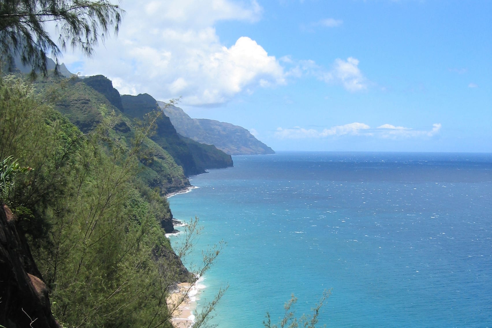 Steep green ridges dropping into a turquoise ocean, with a strip of sandy beach below and white clouds overhead