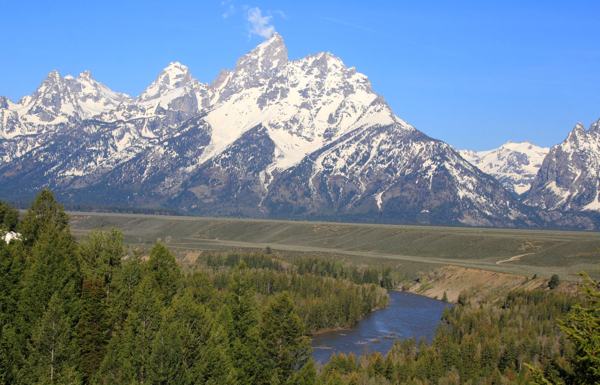 Snow-covered jagged peaks of the Teton Range rising above a forested bend of the Snake River under a clear blue sky