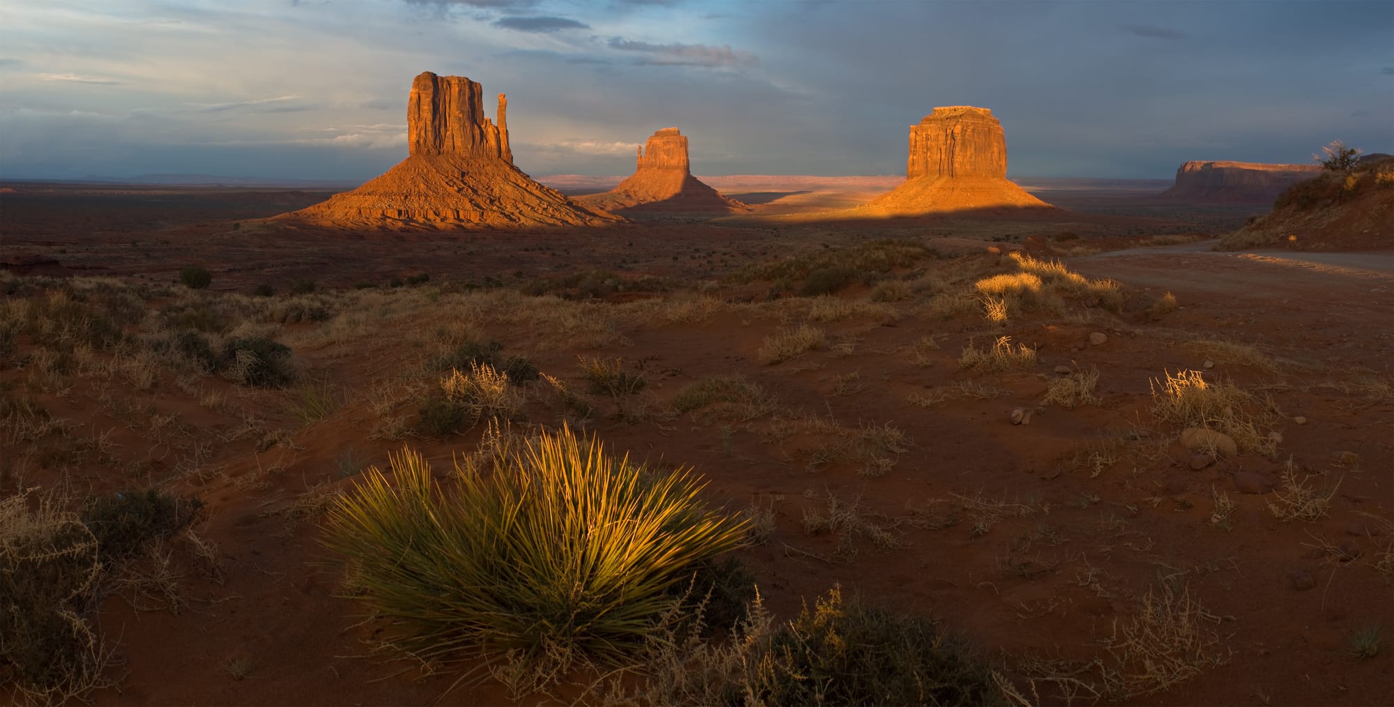 Sandstone buttes glowing orange in low evening sun above a red desert plain dotted with yucca and dry grass
