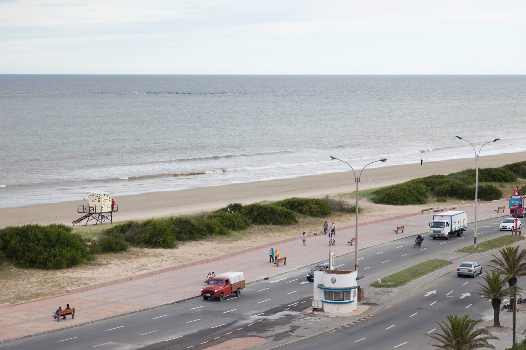 A wide sandy beach and grey water beside a seafront promenade and coastal road, with a lifeguard hut and dunes