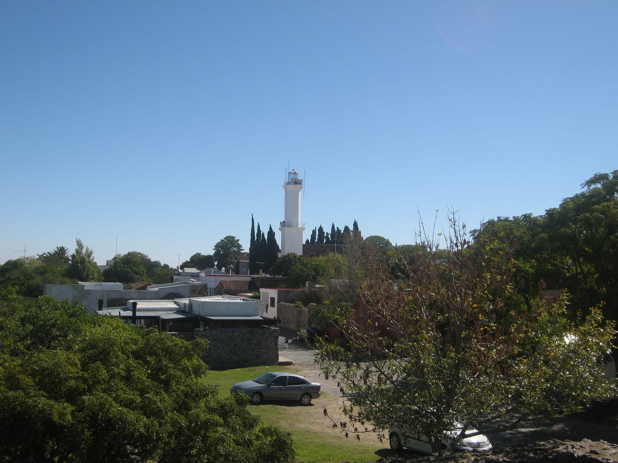 A white stone lighthouse rising above the ruins and red-tiled roofs of an old walled town beside a wide river