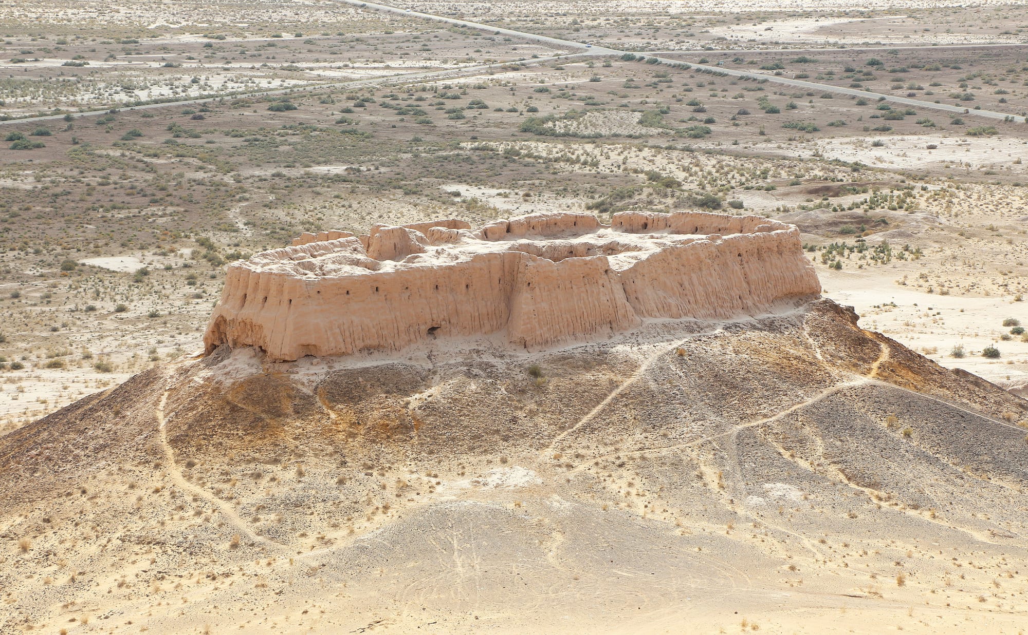 A ruined mud-brick fortress on a bare hillock above a flat desert plain of scrub, salt patches and a straight road