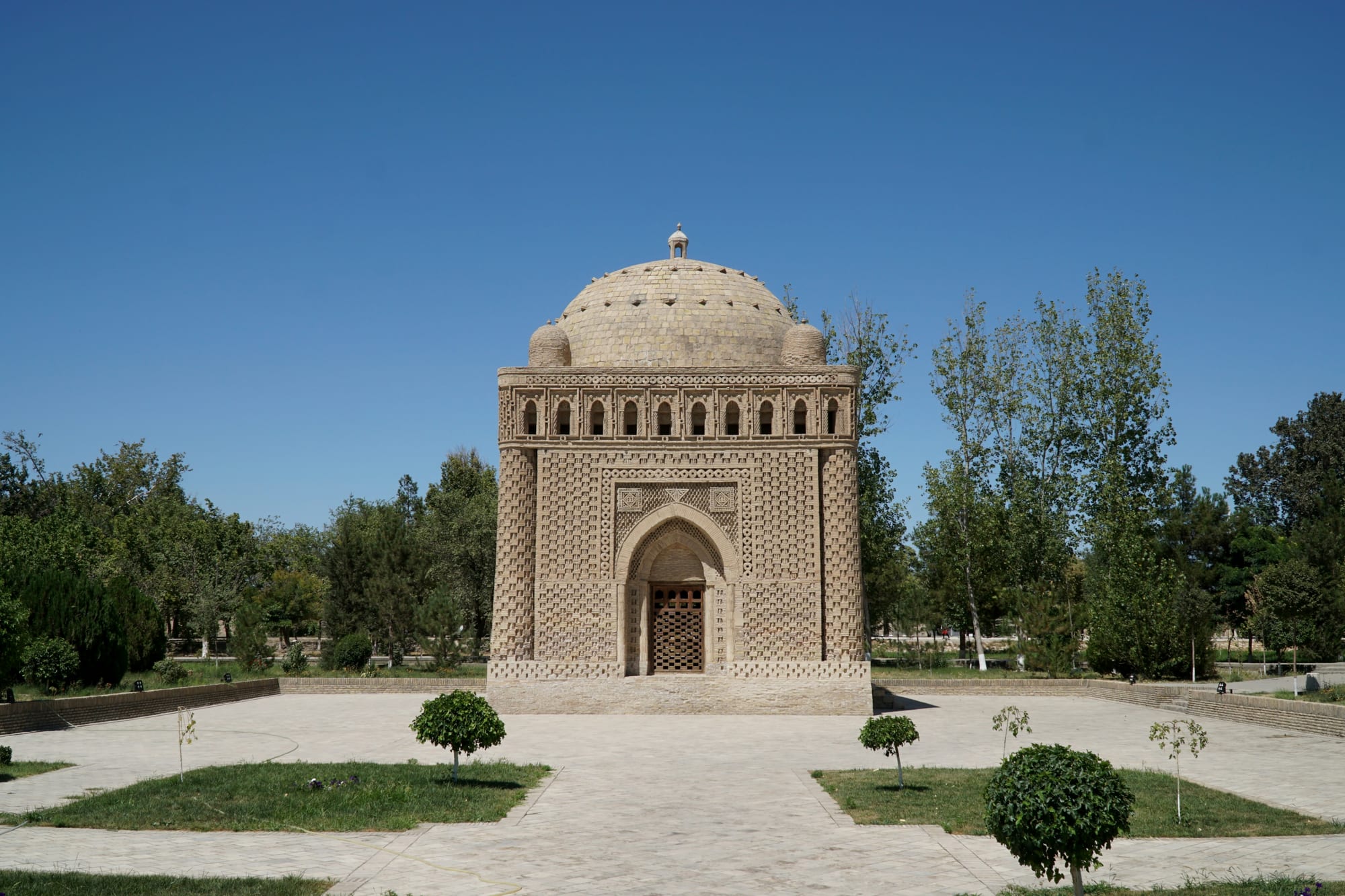 A small cube-shaped domed tomb of patterned tan brickwork standing in a paved park with lawns, young trees and taller poplars under a clear blue sky