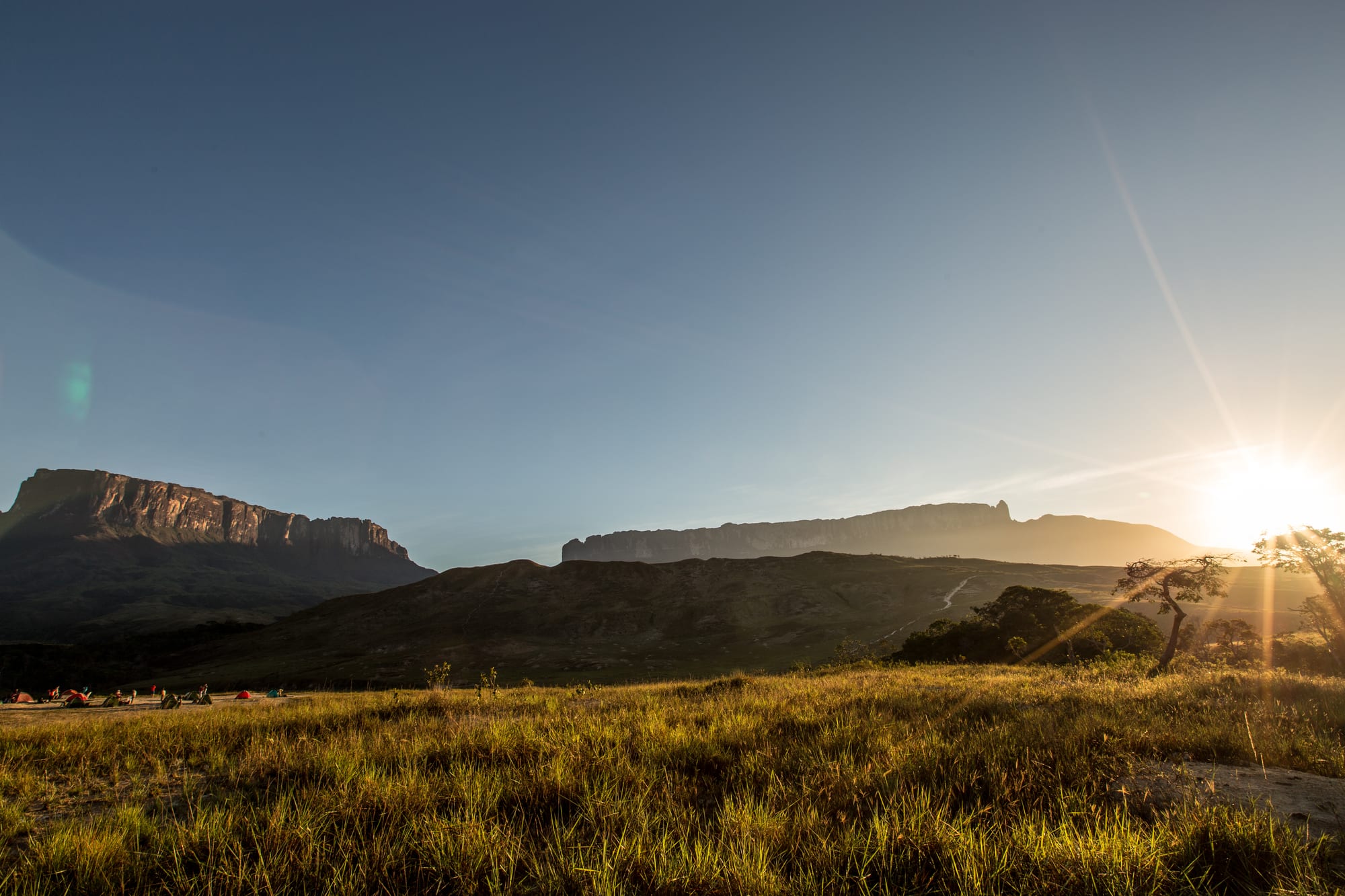Two sheer-walled, flat-topped mountains above golden grassland at sunrise, with small tents in the foreground