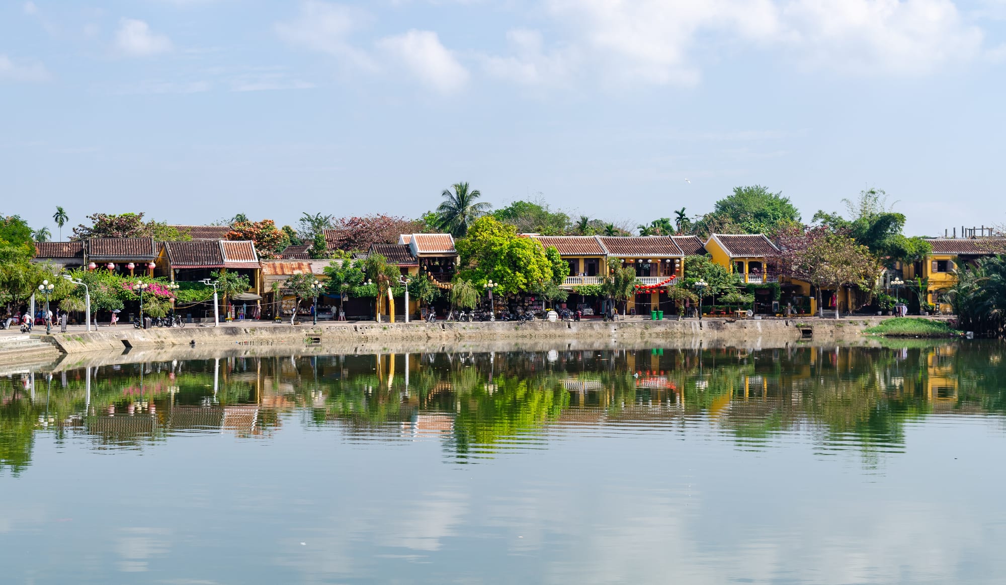 A row of yellow-walled, tile-roofed shophouses among trees along a riverbank, reflected in calm water