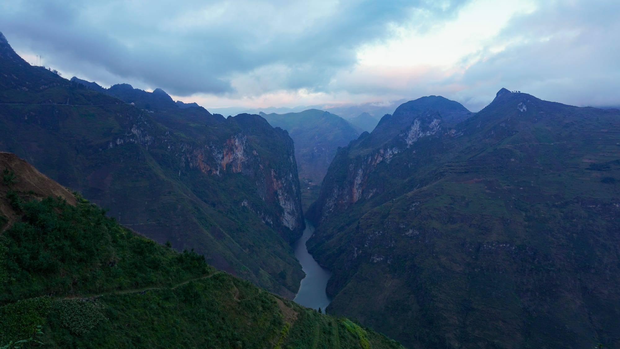 Steep green mountains and grey cliffs falling into a narrow canyon with a river at the bottom under a cloudy sky