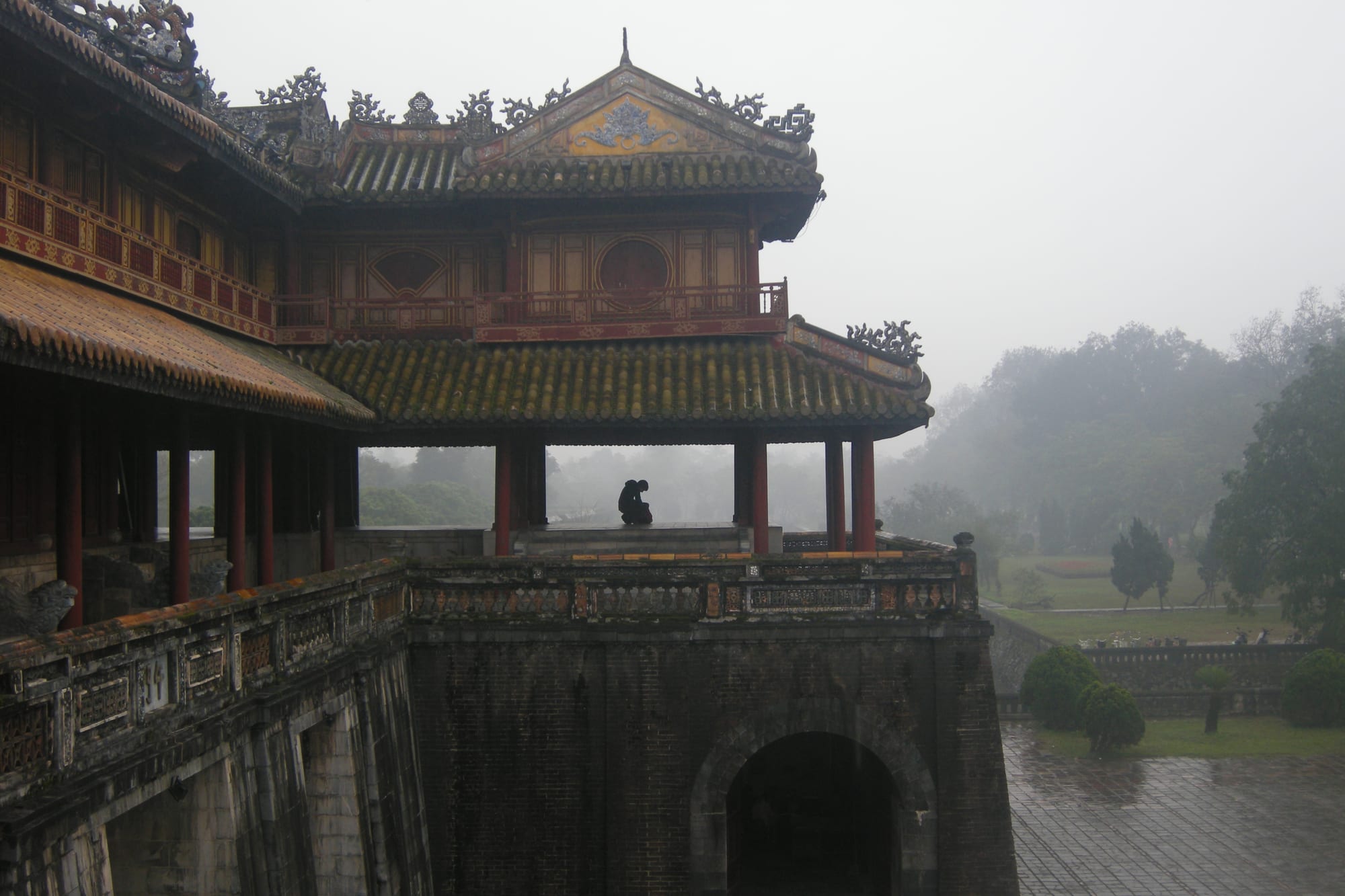 Red-pillared pavilion with tiled roofs on top of a stone gateway, with misty trees and lawns behind