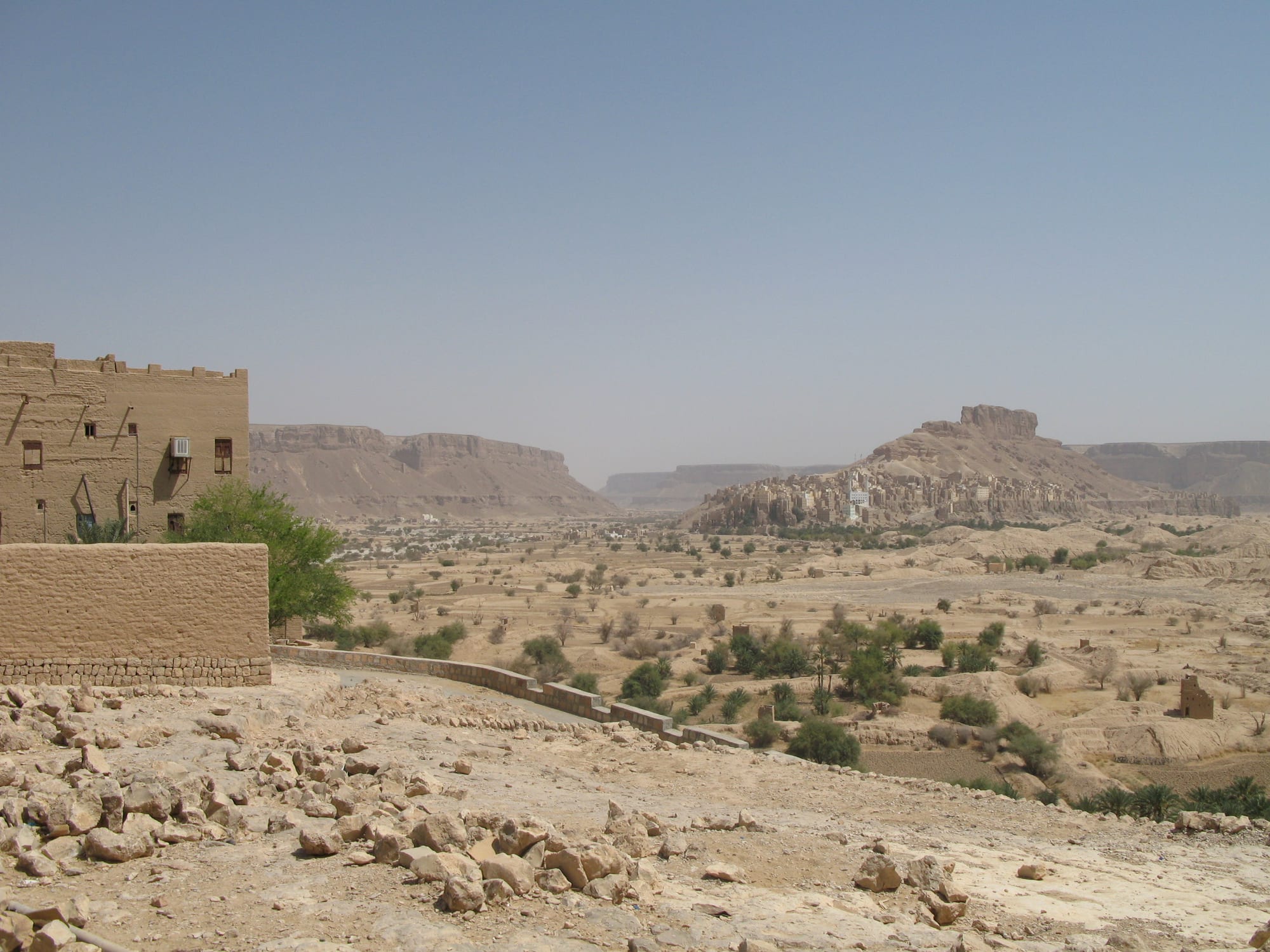 A wide dry valley dotted with green trees and palms, flat-topped cliffs behind, and a mud-brick village climbing a rocky hill on the right