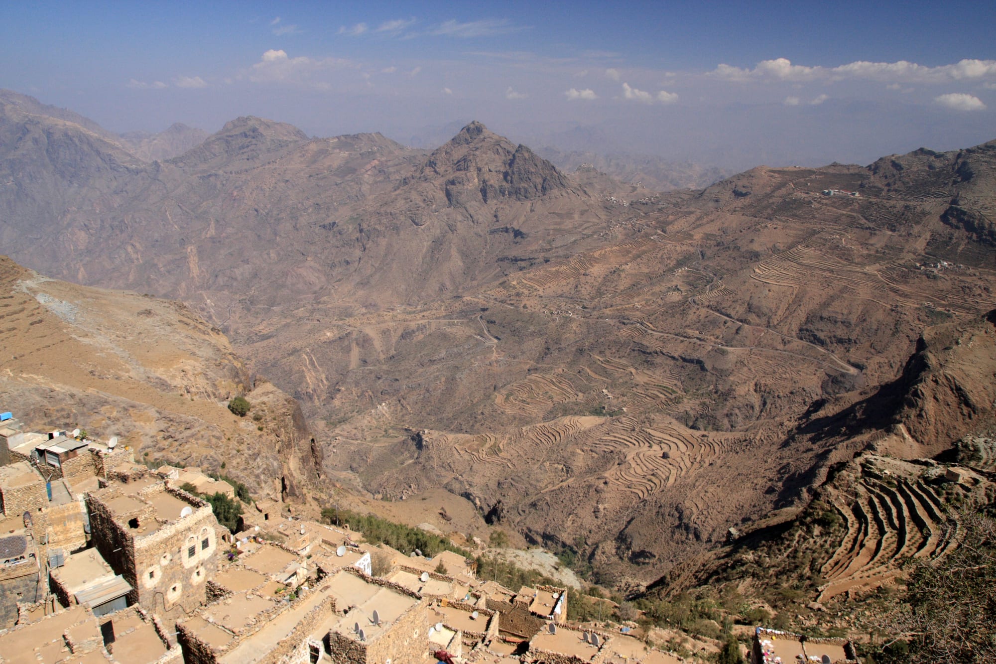 Stone village rooftops on a ridge above a deep mountain valley lined with dry farming terraces and jagged peaks
